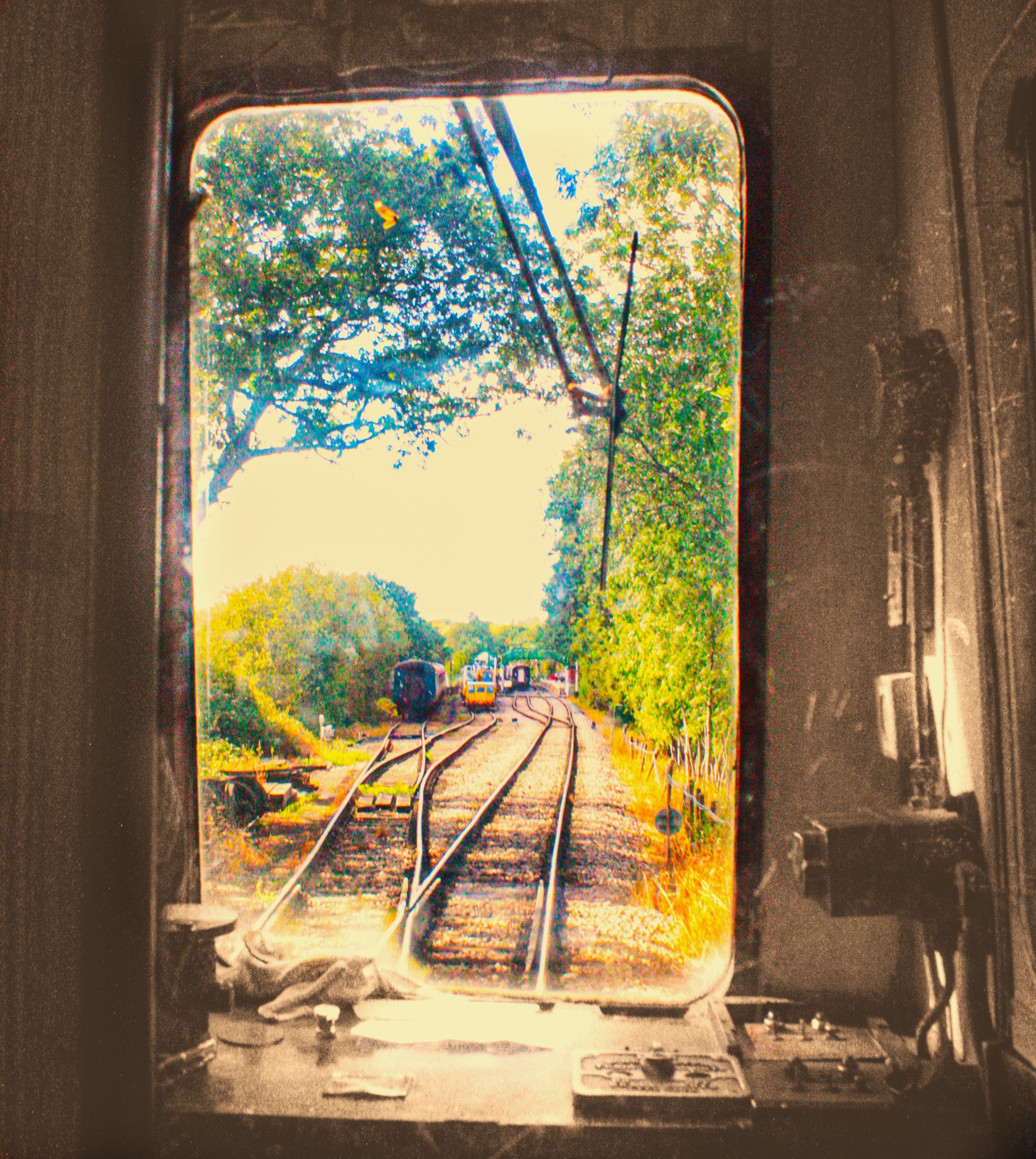 The image shows a scenic view seen from in front of a window, which appears to be part of an old vehicle, possibly a train or bus, with a clear view of a railroad track, trees, and a small white building in the background.
