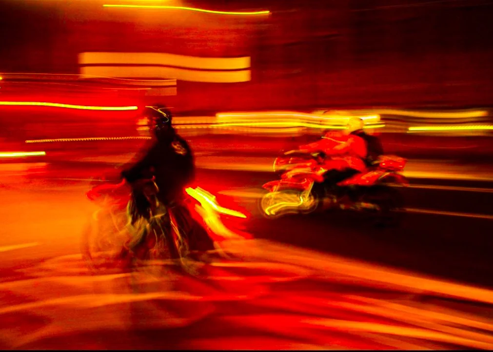 Blurred night scene with two motorcyclists, one wearing a black jacket and helmet, the other in red gear, riding on city streets illuminated by red and yellow lights.