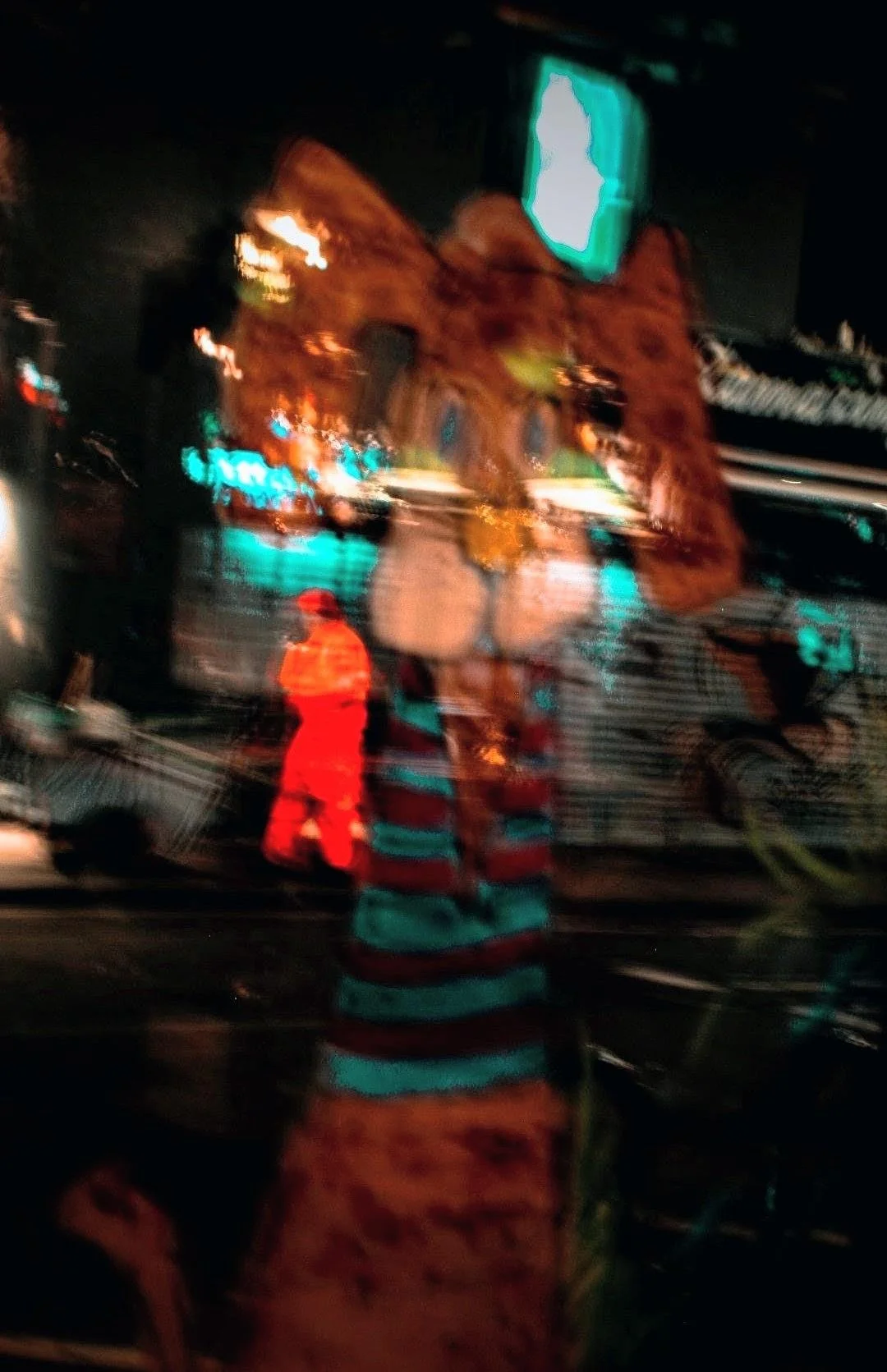 Blurred photo of an outdoor scene at night with a rocky mountain facade in the background, bright neon signs, and people in reflective gear, possibly at a theme park or amusement area.