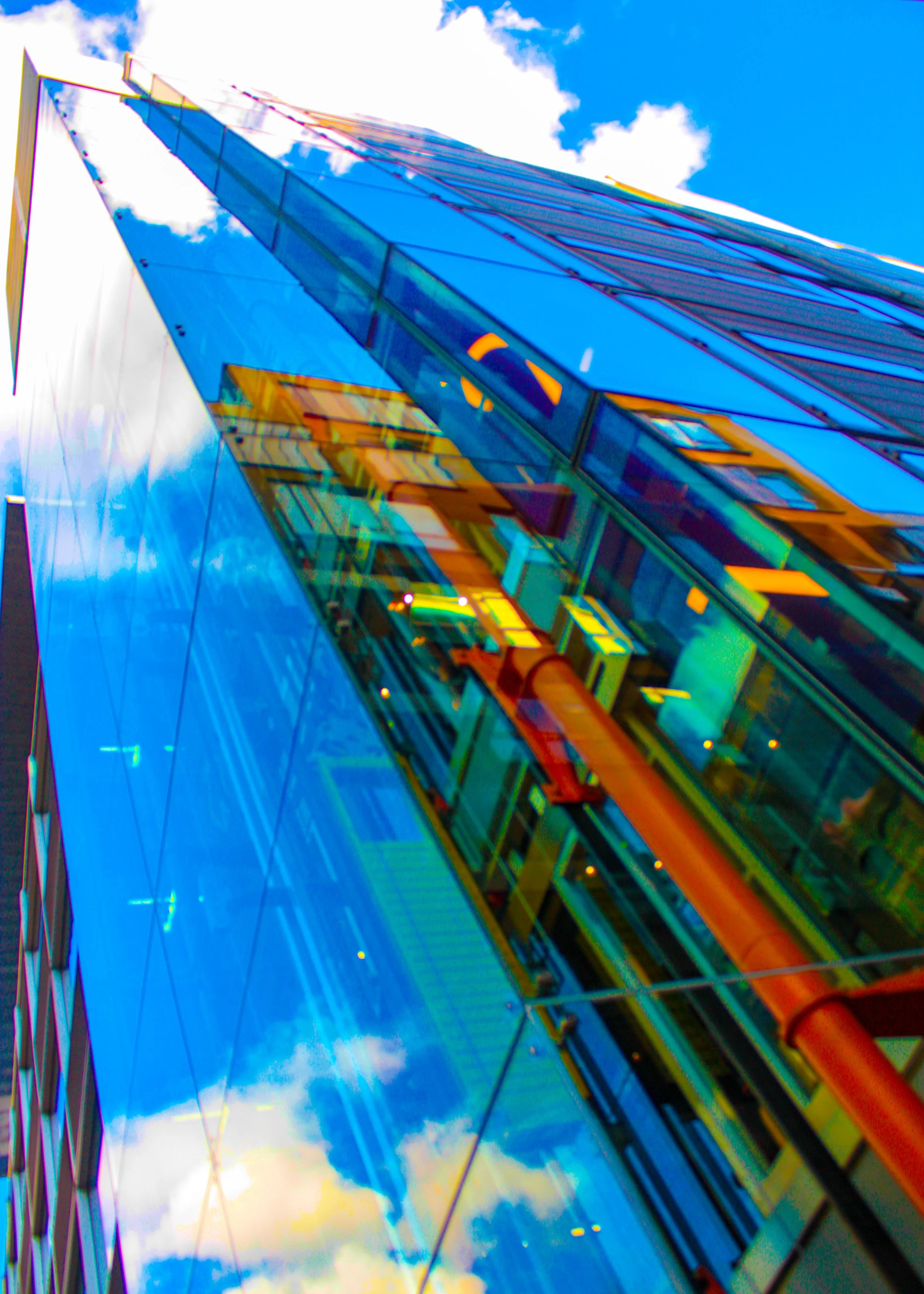 Tall modern glass building reflecting blue sky and clouds, with a visible orange construction pipe and structural elements.