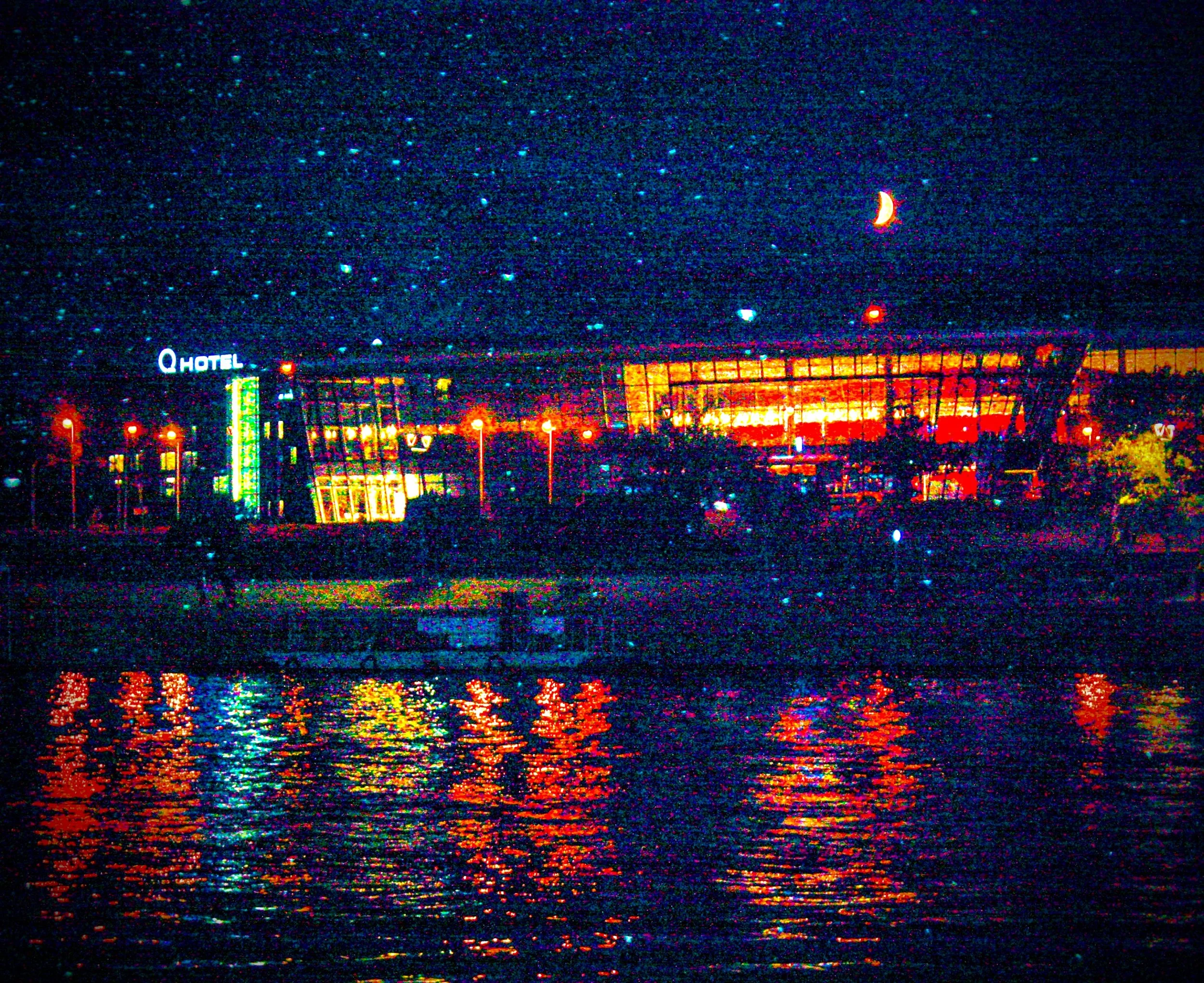 Nighttime cityscape with a starry sky, a crescent moon, and reflections on water, featuring a bright-lit building with a neon sign that reads 'Q Hotel'.