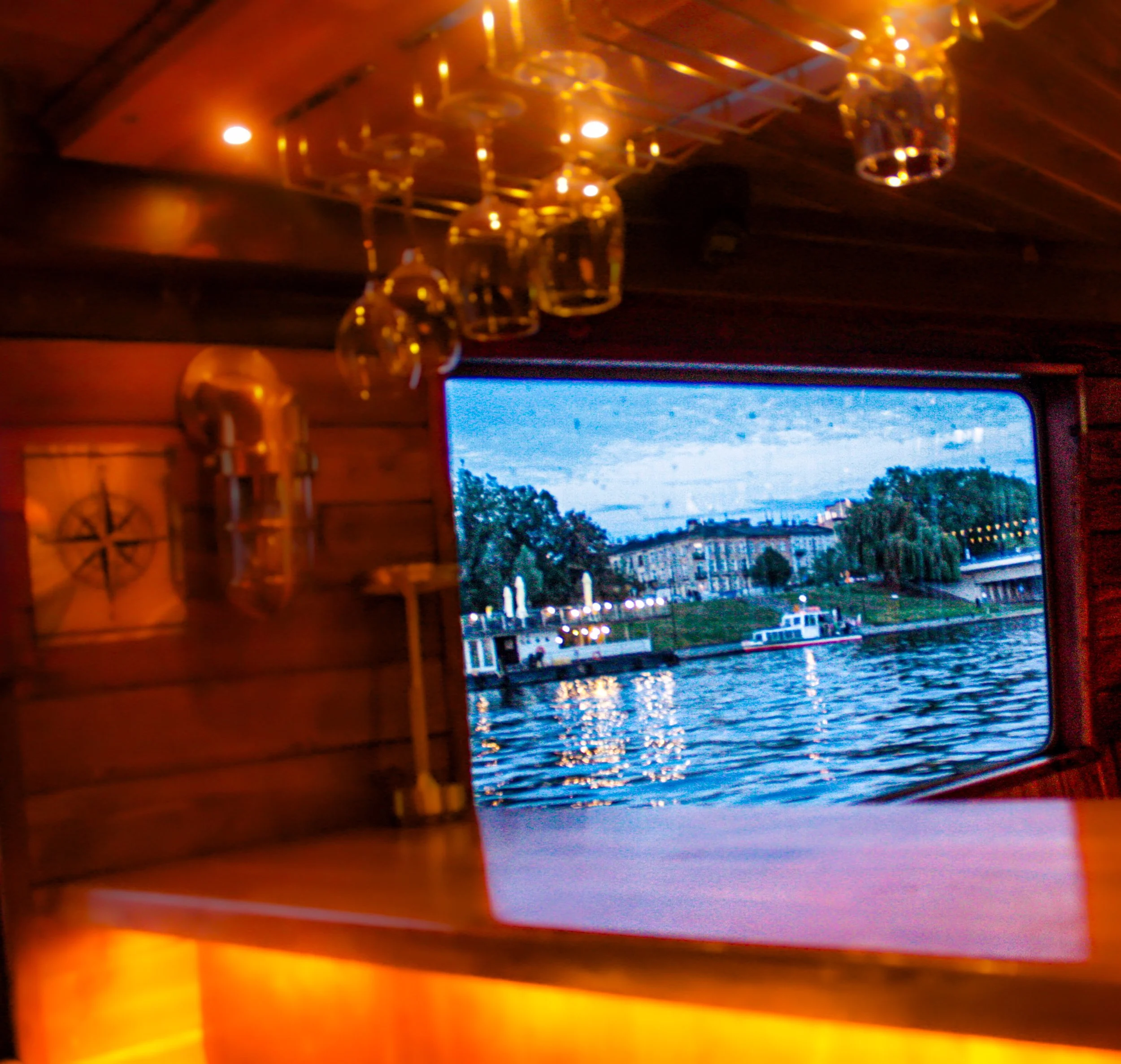 Warm ambient lighting reflecting on glass bottles inside a wooden or brass cabinet, with an outdoor view of water and boats reflecting the evening sky through a window.