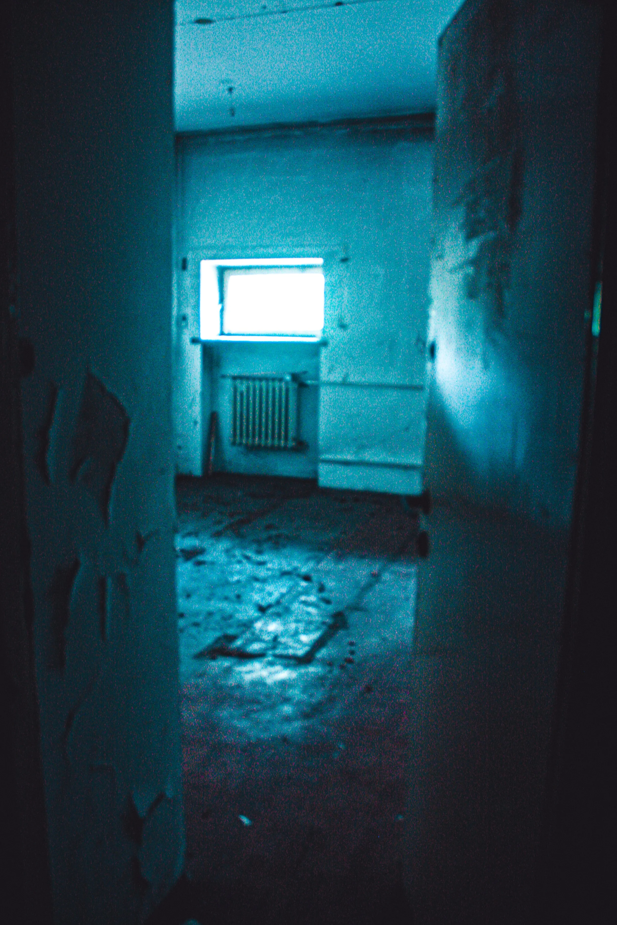Dark, abandoned room with a small window emitting bright light, a radiator below the window, and debris on the floor. The walls are peeling and the space appears neglected.