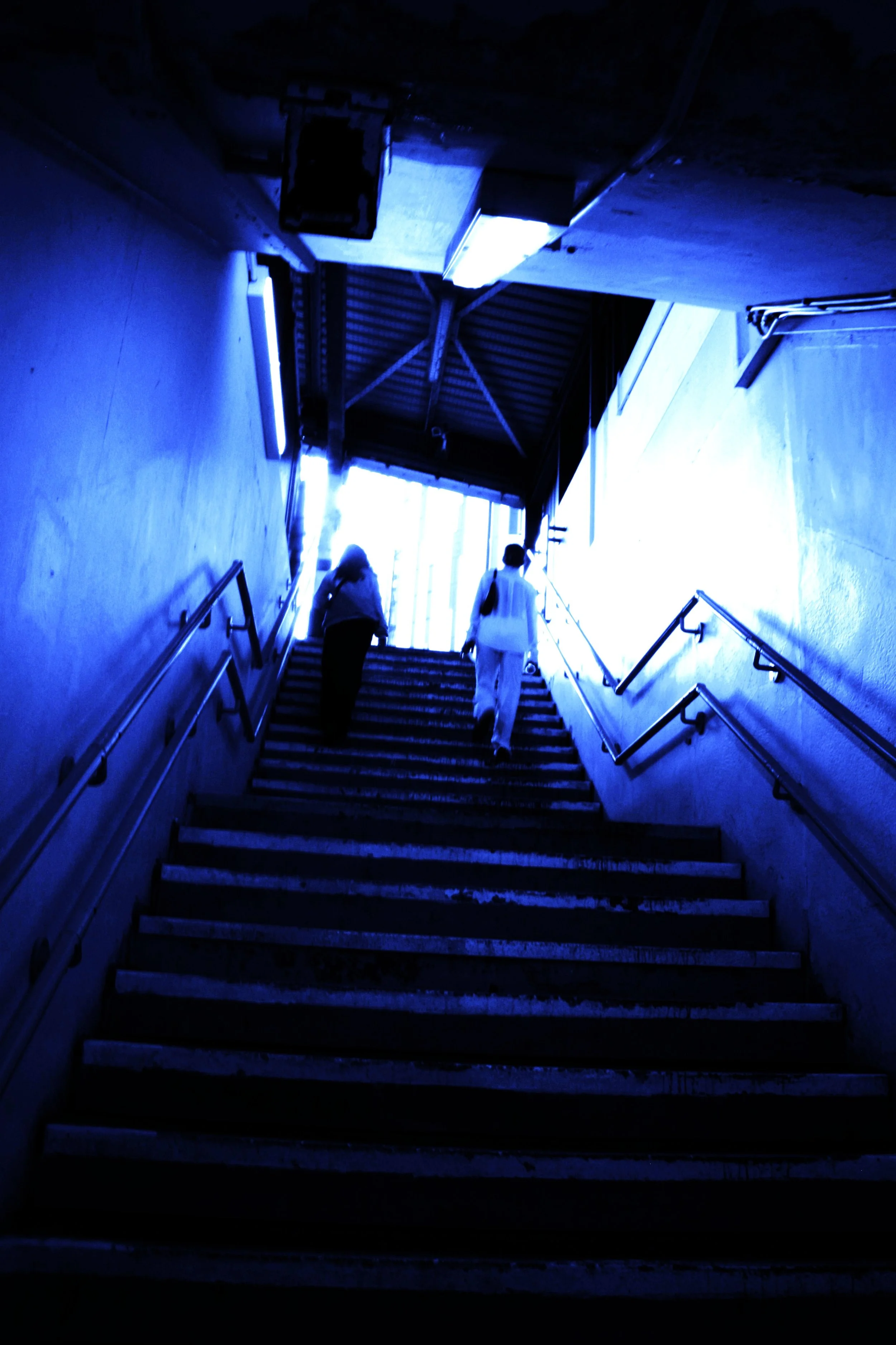 A staircase with blue-tinted lighting, three people walking up, and a covered entrance at the top with a corrugated metal roof.