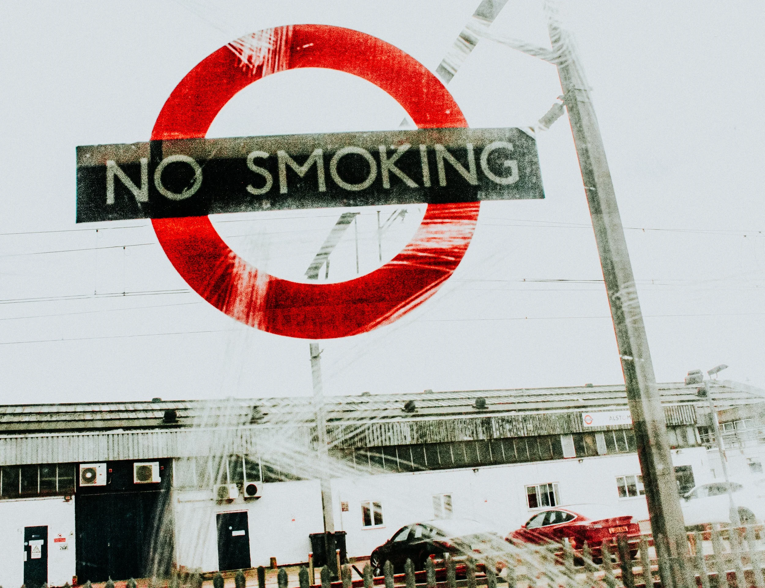 London Underground 'No Smoking' sign on a pole against a cloudy sky, with parking lot and building in the background.