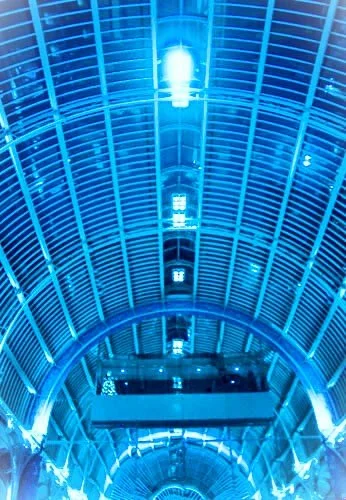 Inside a modern building with brightly lit blue ceiling and glass walls, decorated with lights and architectural details.