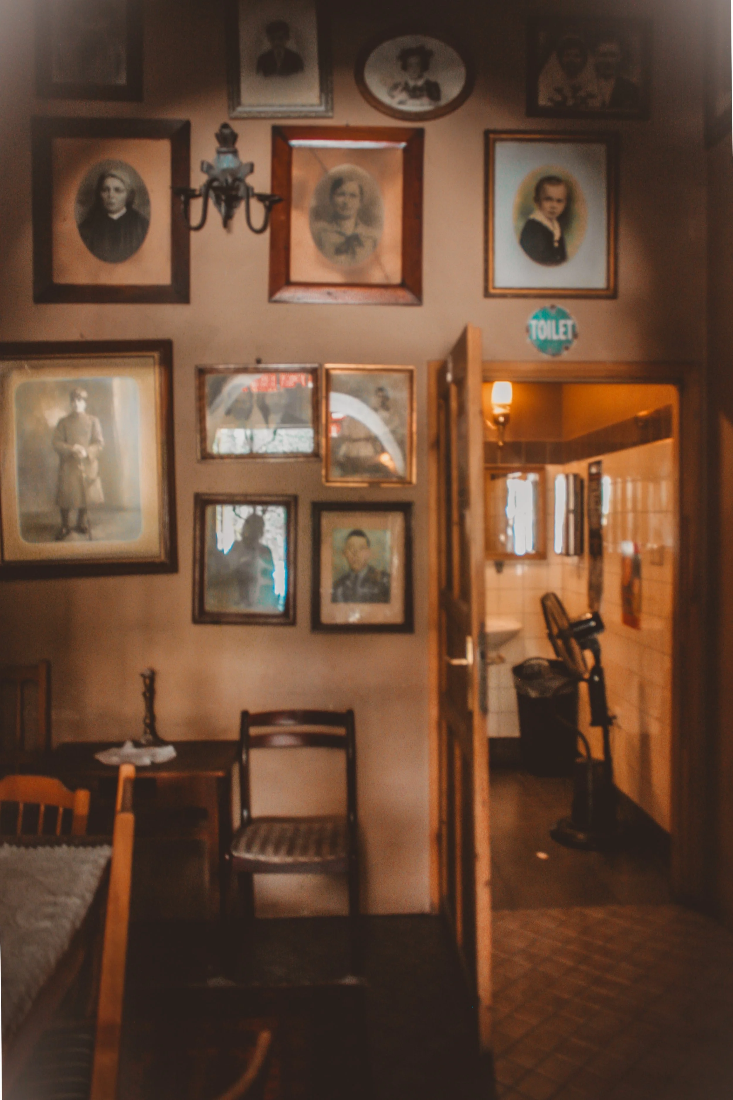 A cozy, vintage-style interior with framed black-and-white photographs on the wall, wooden furniture including chairs and a table, a small chandelier, a sign for the toilet, and a glimpse of a kitchen area with tiled flooring.