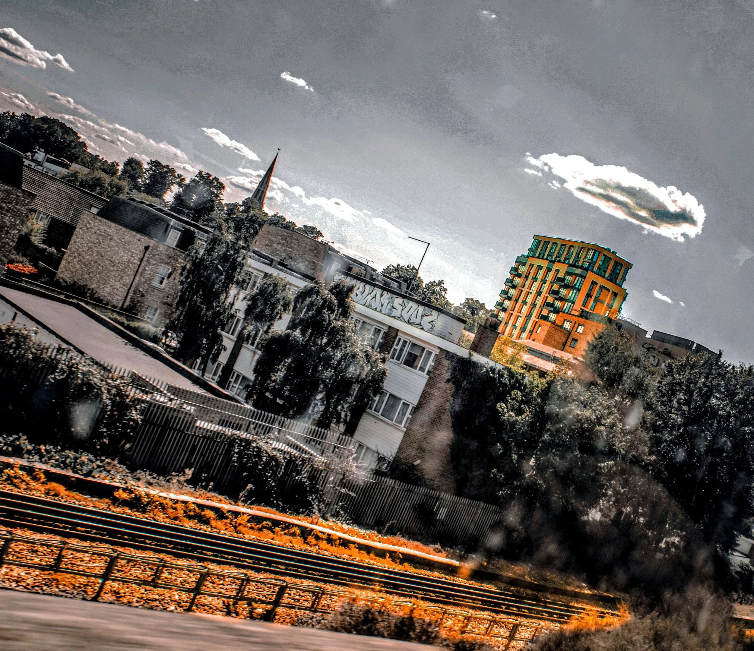 A cityscape showing modern buildings, a church steeple, trees, and a cloudy sky, with a metal railing in the foreground.