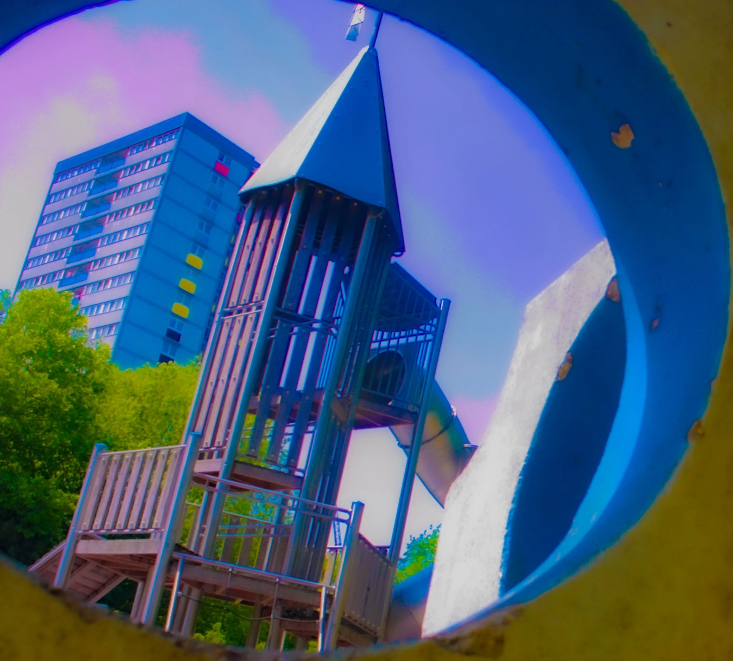 Looking through a circular hole in a concrete structure, there is a modern building with multiple balconies and colorful window accents, set against a blue sky with some trees in the foreground.