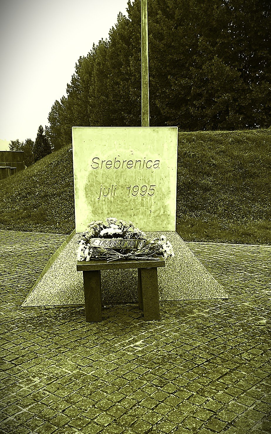 Memorial with flowers and a sign reading 'Srebrenica July 1995' in front of a grassy hill and trees.