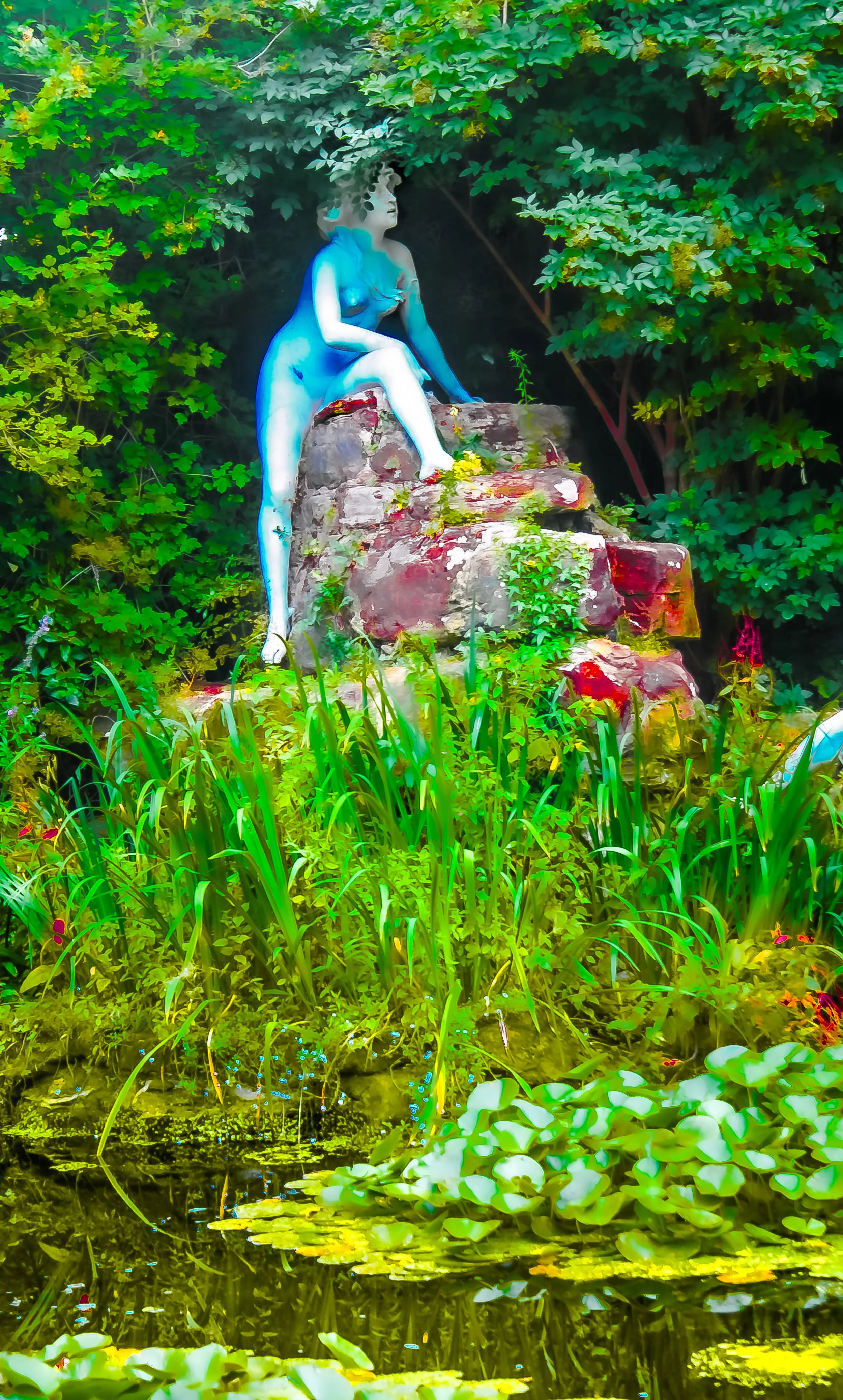 A statue of a woman sitting on a rock, surrounded by lush green trees and plants, with a pond below reflecting the greenery.