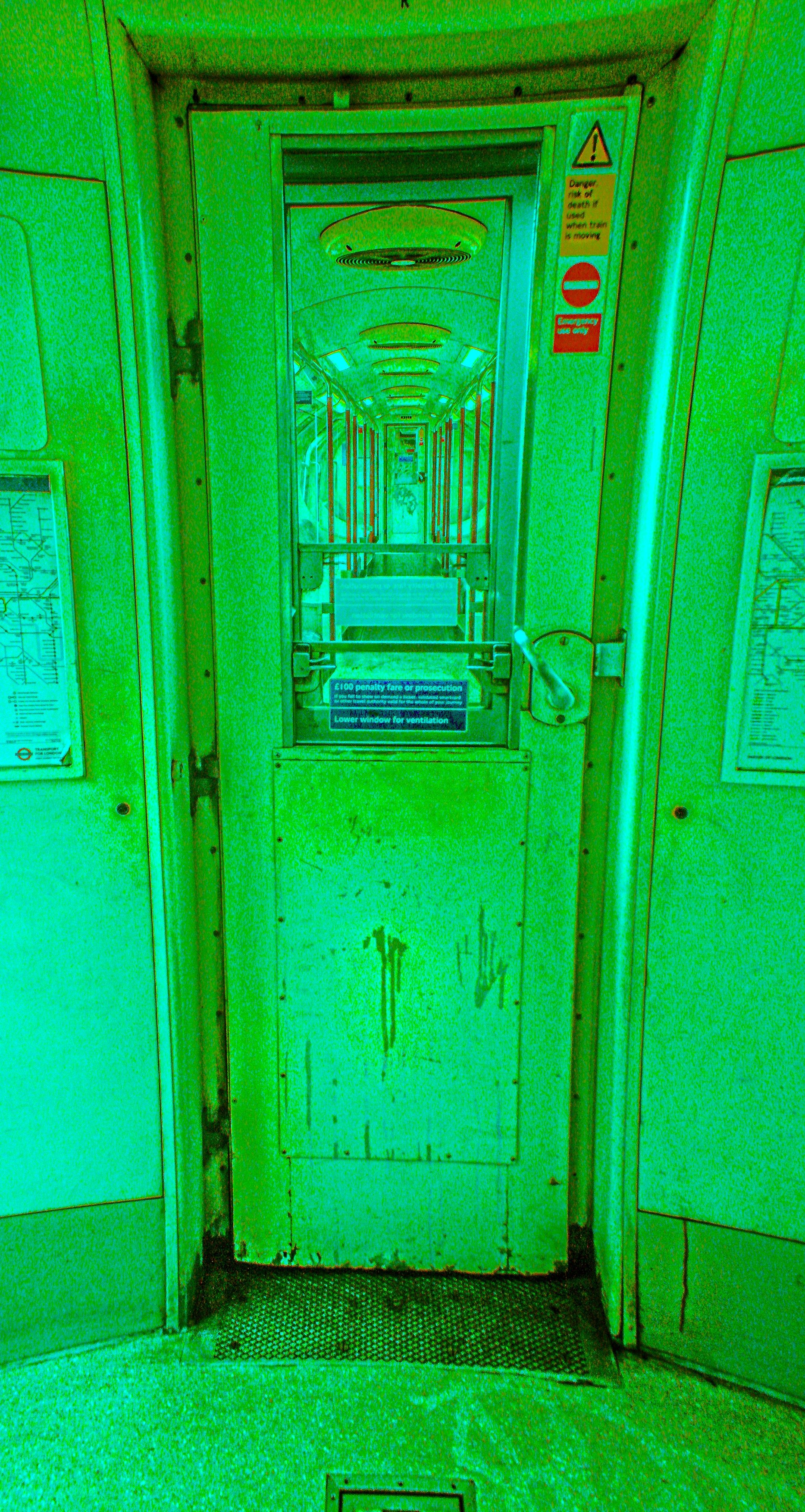 A metal gate with a sign that reads '£100 penalty fare or prosecution' in a tunnel or enclosed walkway, flanked by walls with maps and notices.