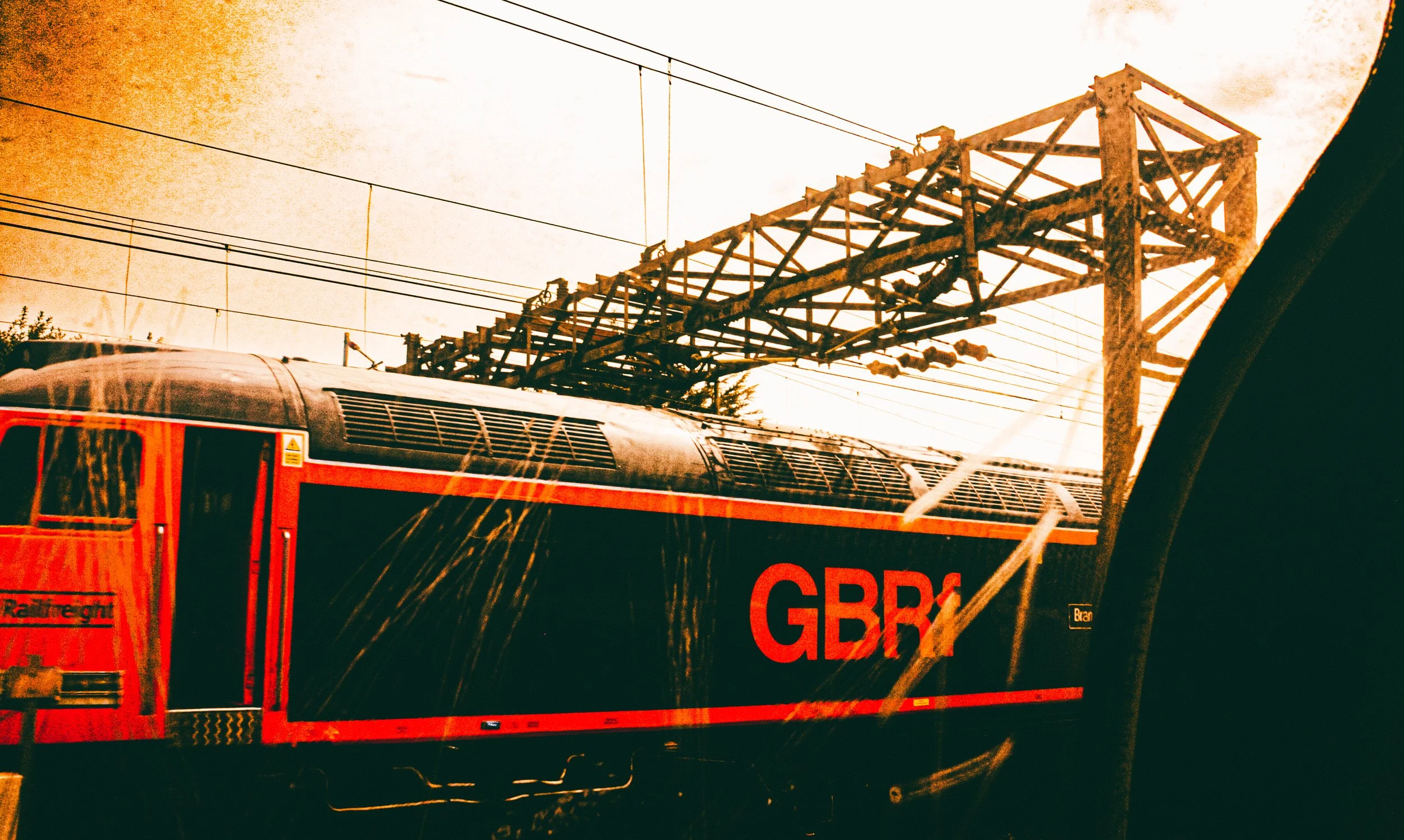 Part of a red and black train with the letters 'GBX' on the side, passing under a large metal railway bridge with electrical wires overhead.