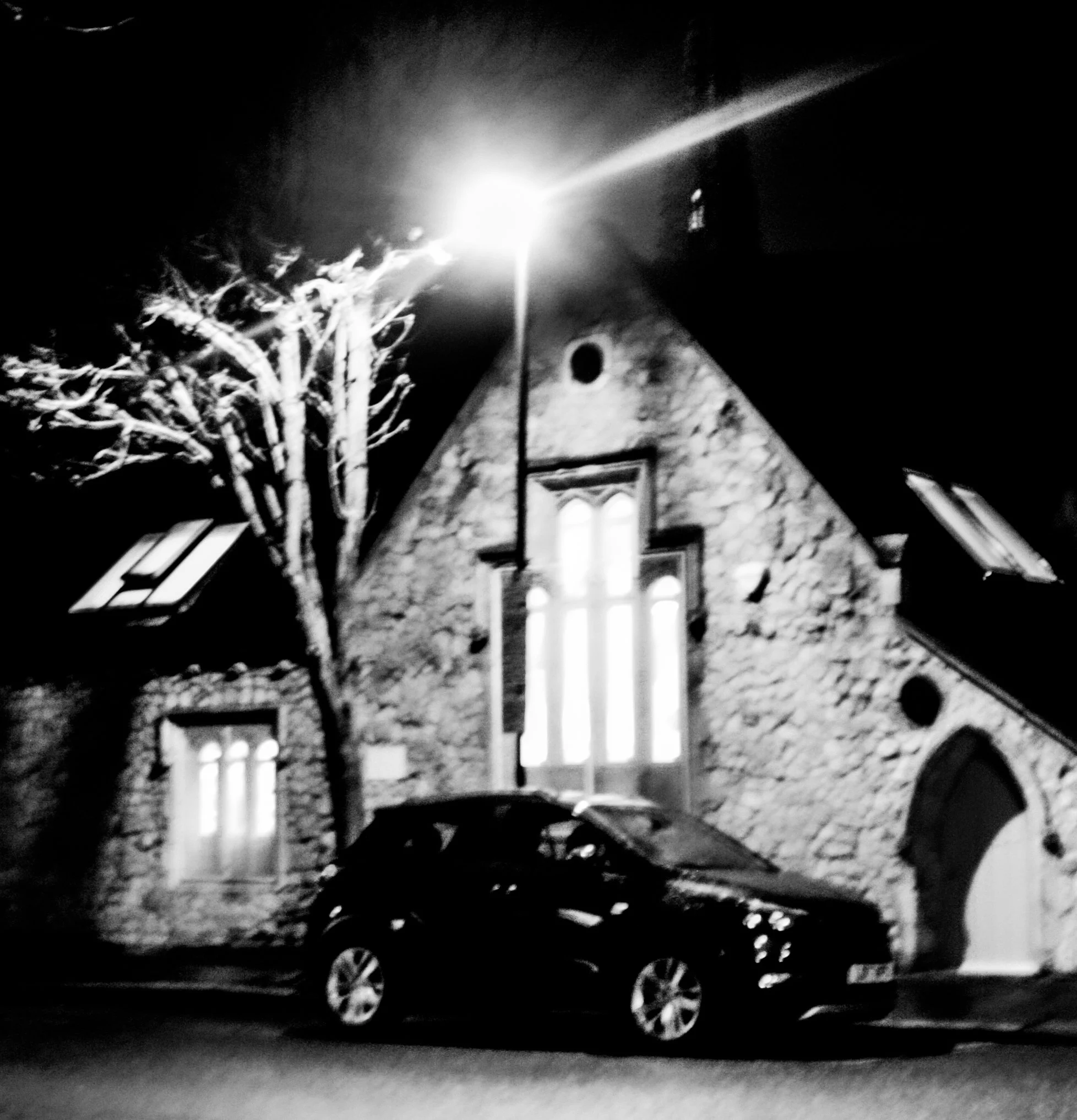 Black and white photo of a house with a steep roof, illuminated by a bright streetlamp, a barren tree in the foreground, and parked cars along the street at night.