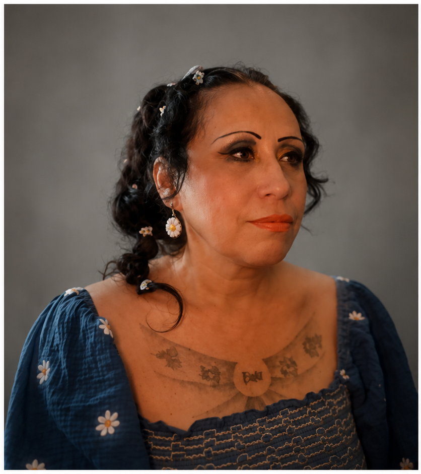 A woman with dark, curly hair styled with small daisy hair accessories, wearing makeup with prominent eyeliner and orange lipstick, and daisy earrings. She's dressed in a blue dress with daisy embroidery and a sheer top with a butterfly necklace. The background is plain and gray.