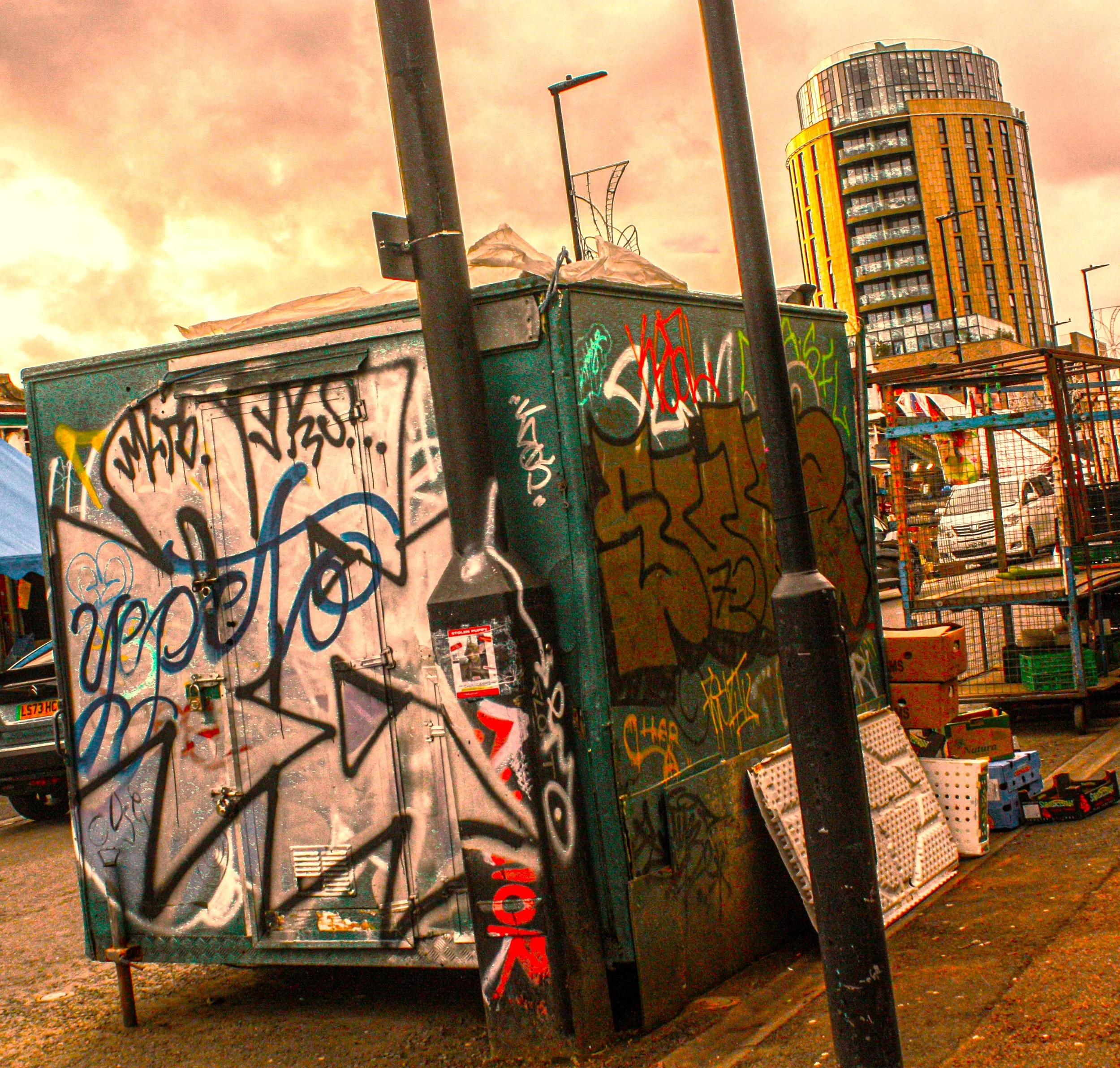 Graffiti-covered metal structures in an urban setting with a tall building in the background and a cloudy sky.