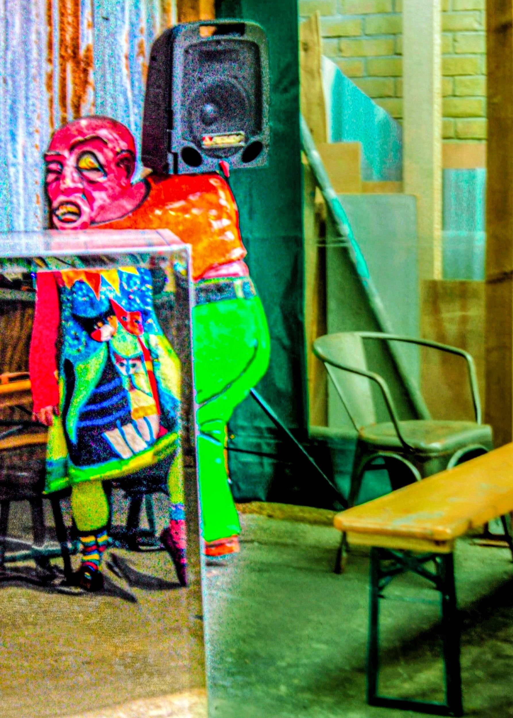 A colorful clown or performer costume with a red face mask, rainbow socks, and a multicolored outfit displayed on a stand. In the background, there is a black speaker, a green chair, a beige chair, a table, and a staircase with yellow and turquoise decorations.