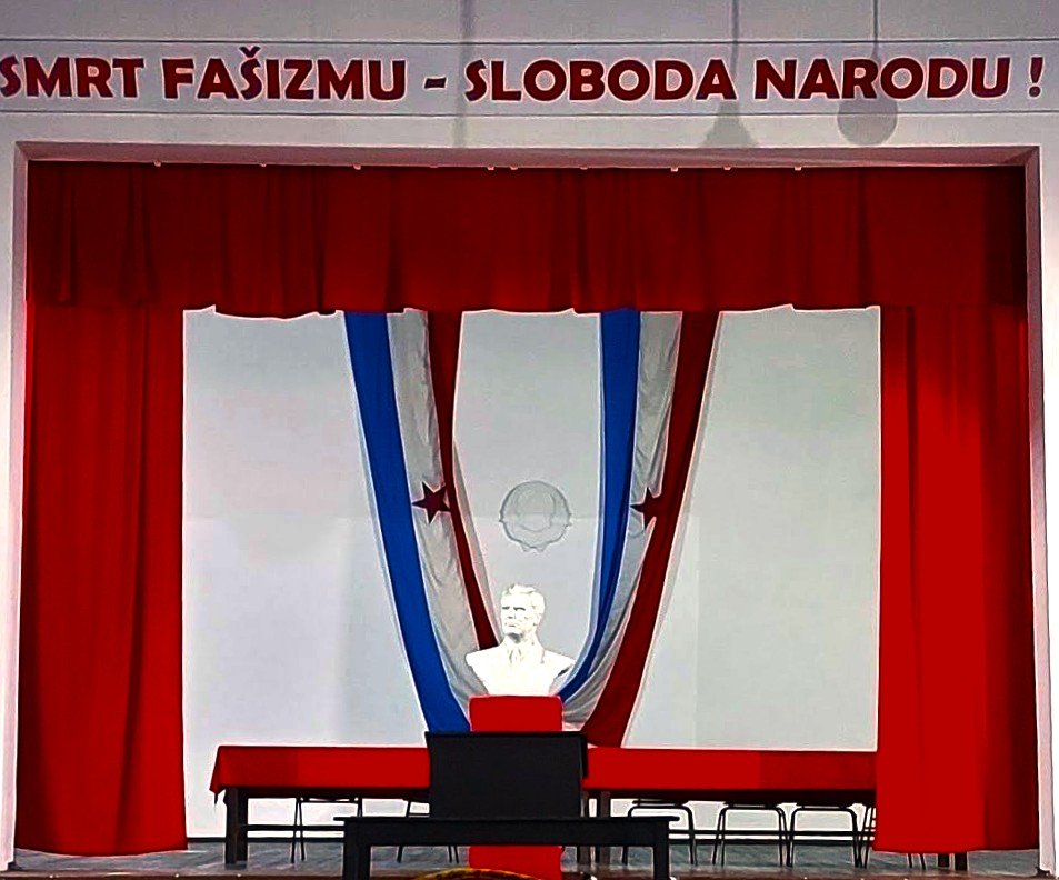 Stage with red curtains, a statue of a man in front of a backdrop of blue, white, and red drapes, and a banner with text in a Slavic language.
