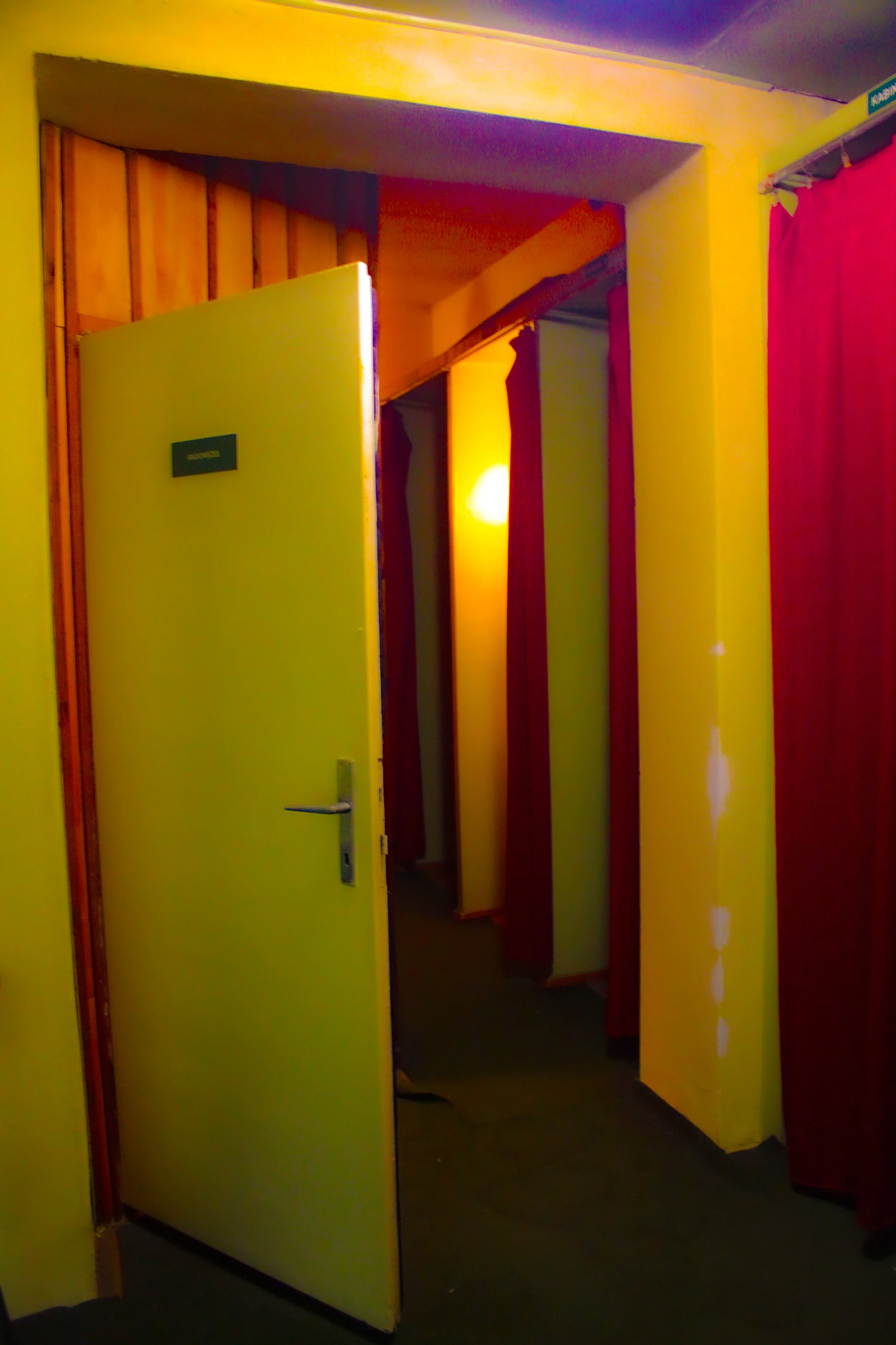 A partially opened door with a sign indicating it is a shower, revealing a dimly lit hallway with yellow walls and red curtains.