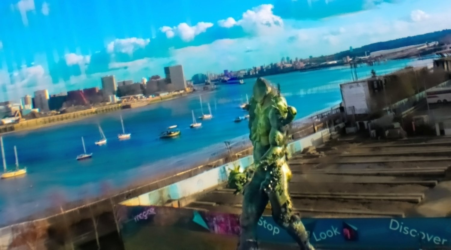 View of a river with sailboats, a city skyline with tall buildings, and a statue in the foreground on a sunny day.