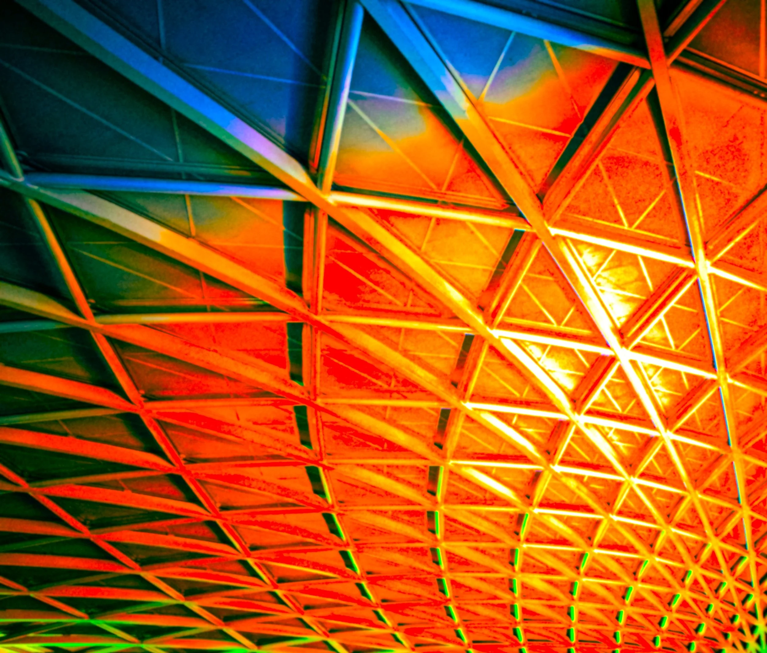 Close-up of a colorful geometric ceiling with illuminated triangular metal framework, featuring vibrant orange, yellow, blue, and green hues.