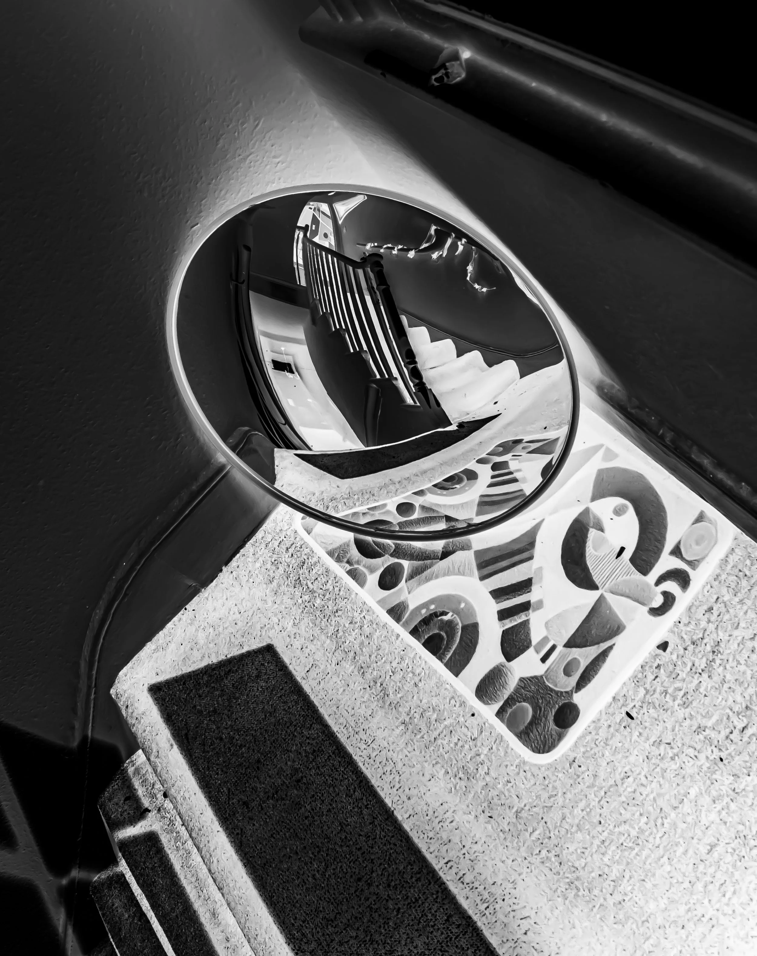 Close-up of a staircase viewed through a circular mirror, with a decorative patterned mat on the floor. The photo is in black and white.