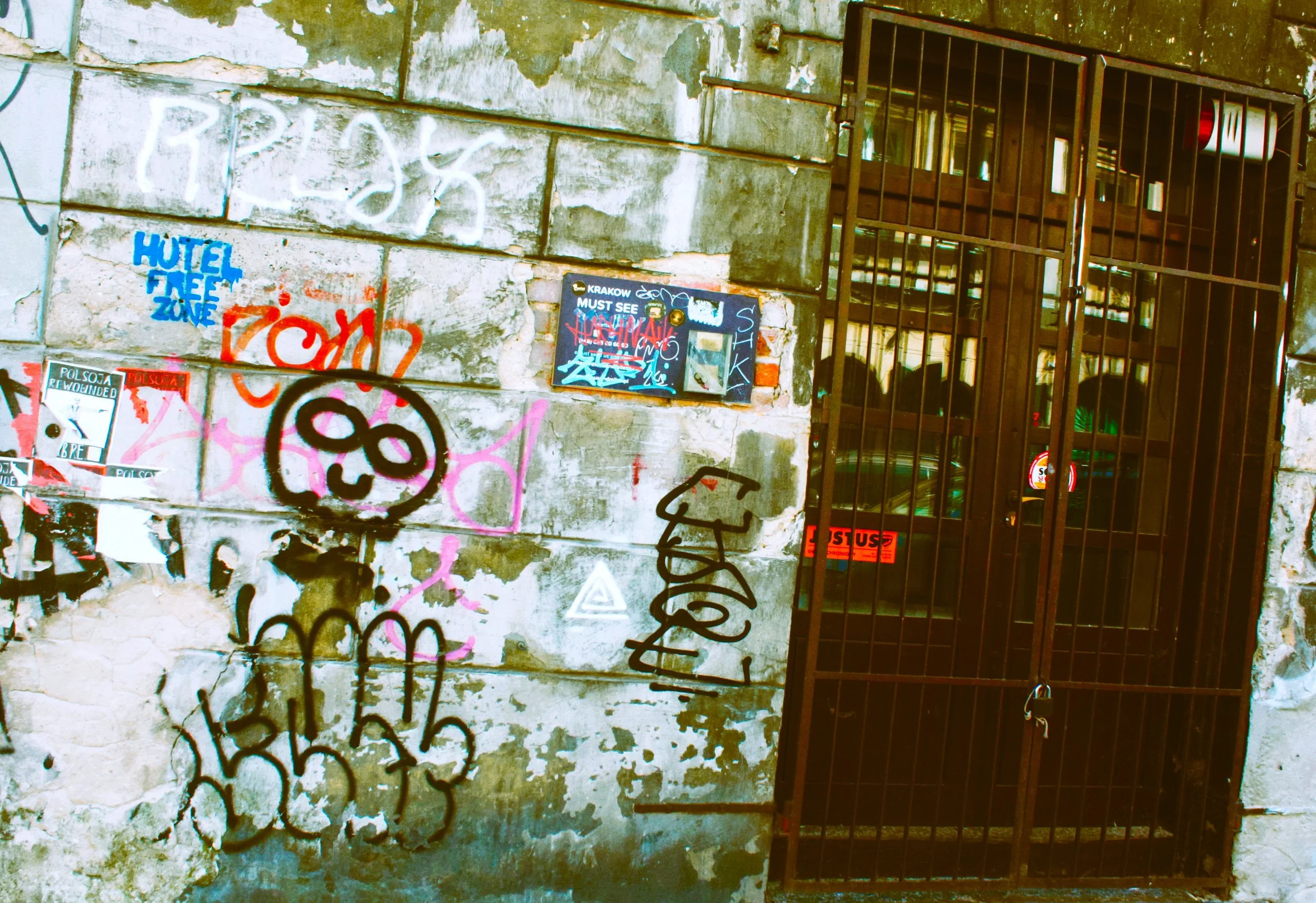 A worn, graffiti-covered wall with a metal gate, some spray paint tags, stickers, and a small sign that says "KRAKOW MUST SEE".