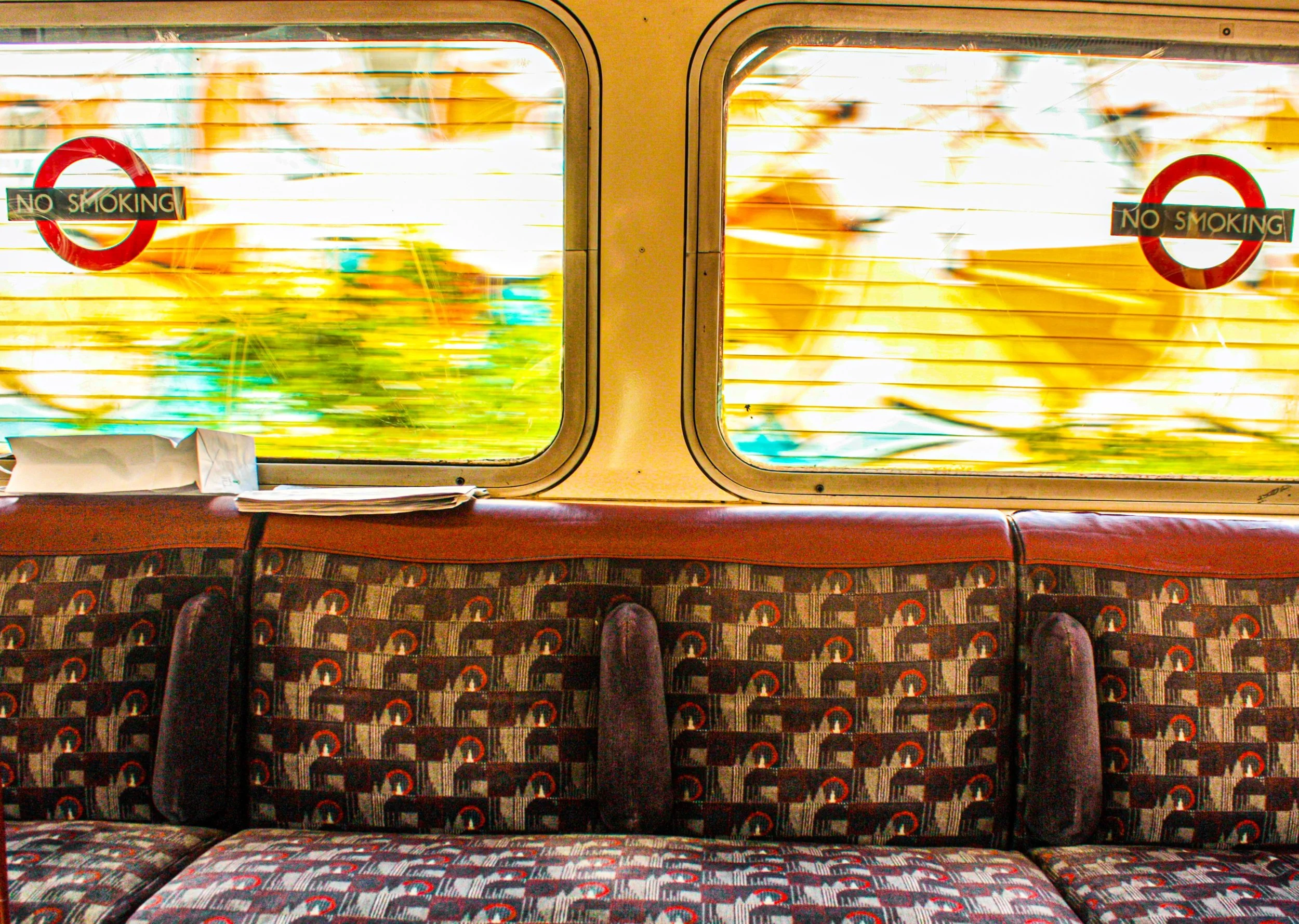 Inside a train, view of two rectangular windows with blinds, displaying blurred greenery and outdoor scenery, and signs reading "No Smoking" on each window. The seats below are arranged in rows with patterned upholstery, and some personal belongings are visible on the seats.