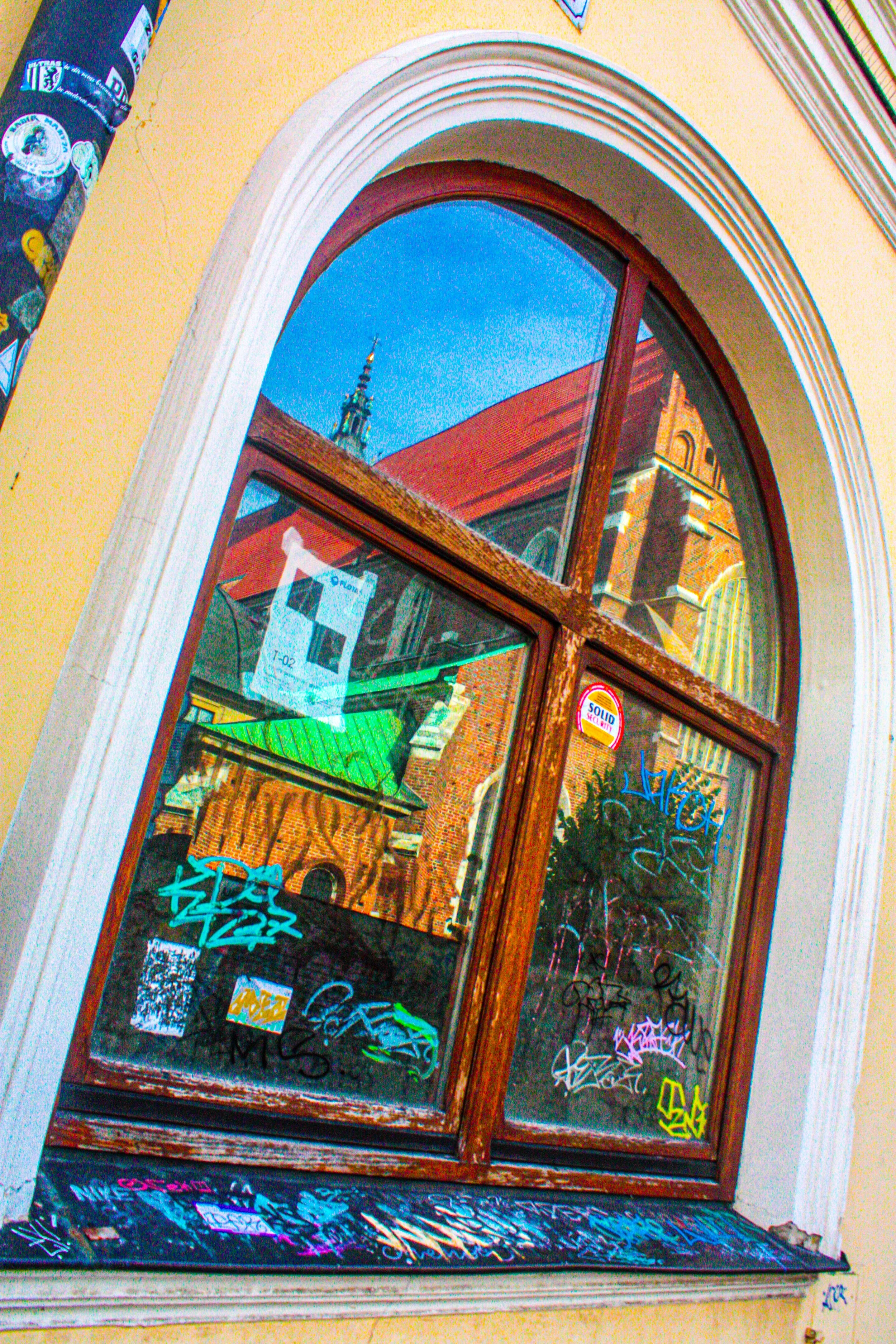 A window with graffiti and stickers on the glass, reflecting a church steeple and a red brick building with arched windows and decorative brickwork.