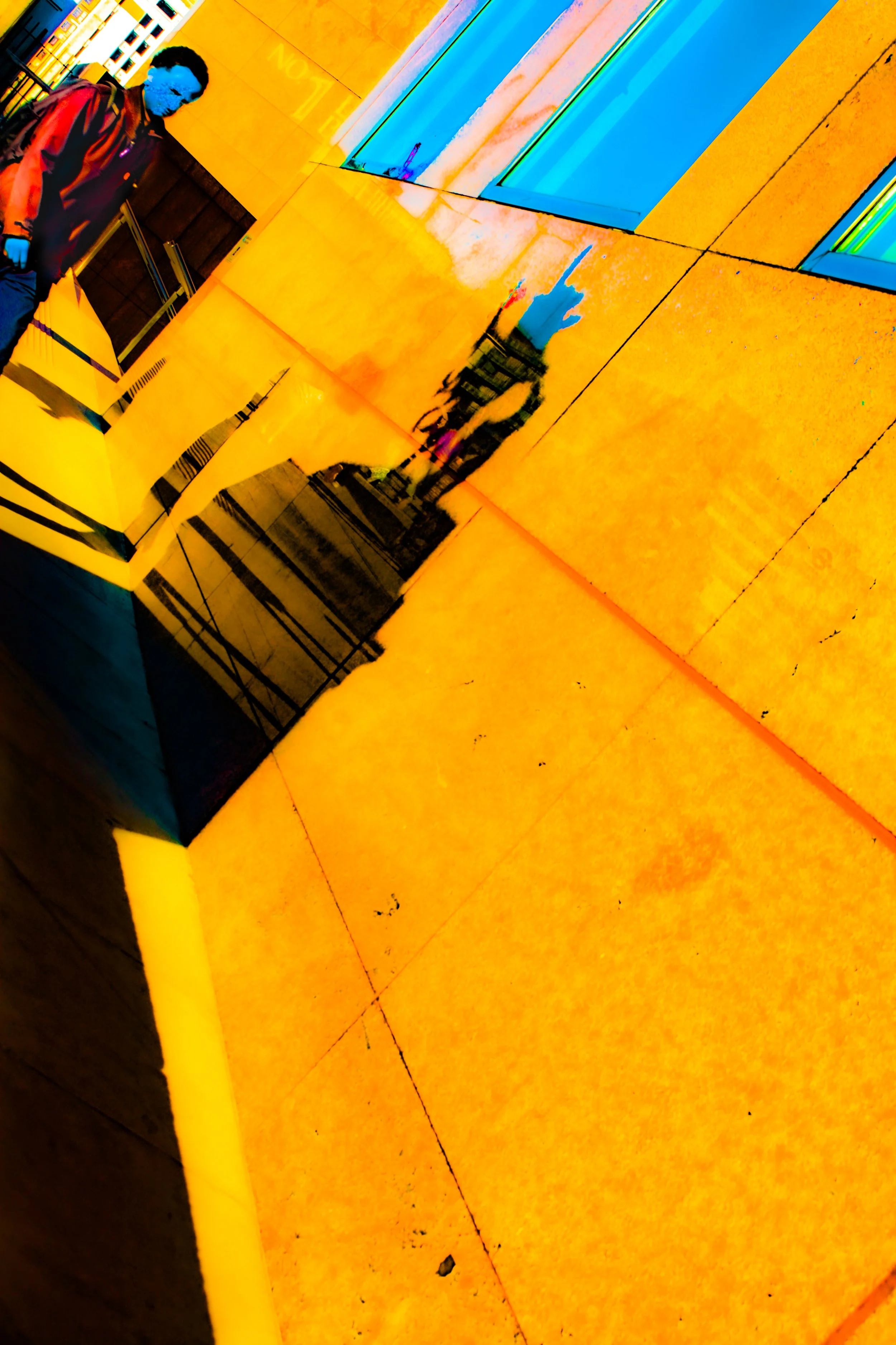 A person with a blue face and hands, wearing a red and black jacket, standing near a yellow textured wall or structure with reflections of other people and buildings in shiny surfaces.