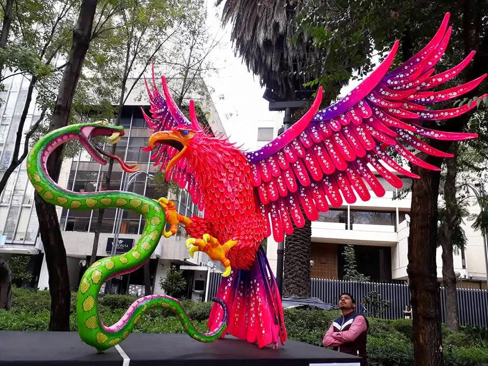 Colorful sculpture of a serpent and a phoenix on display outdoors in an urban setting, with trees and modern buildings in the background.