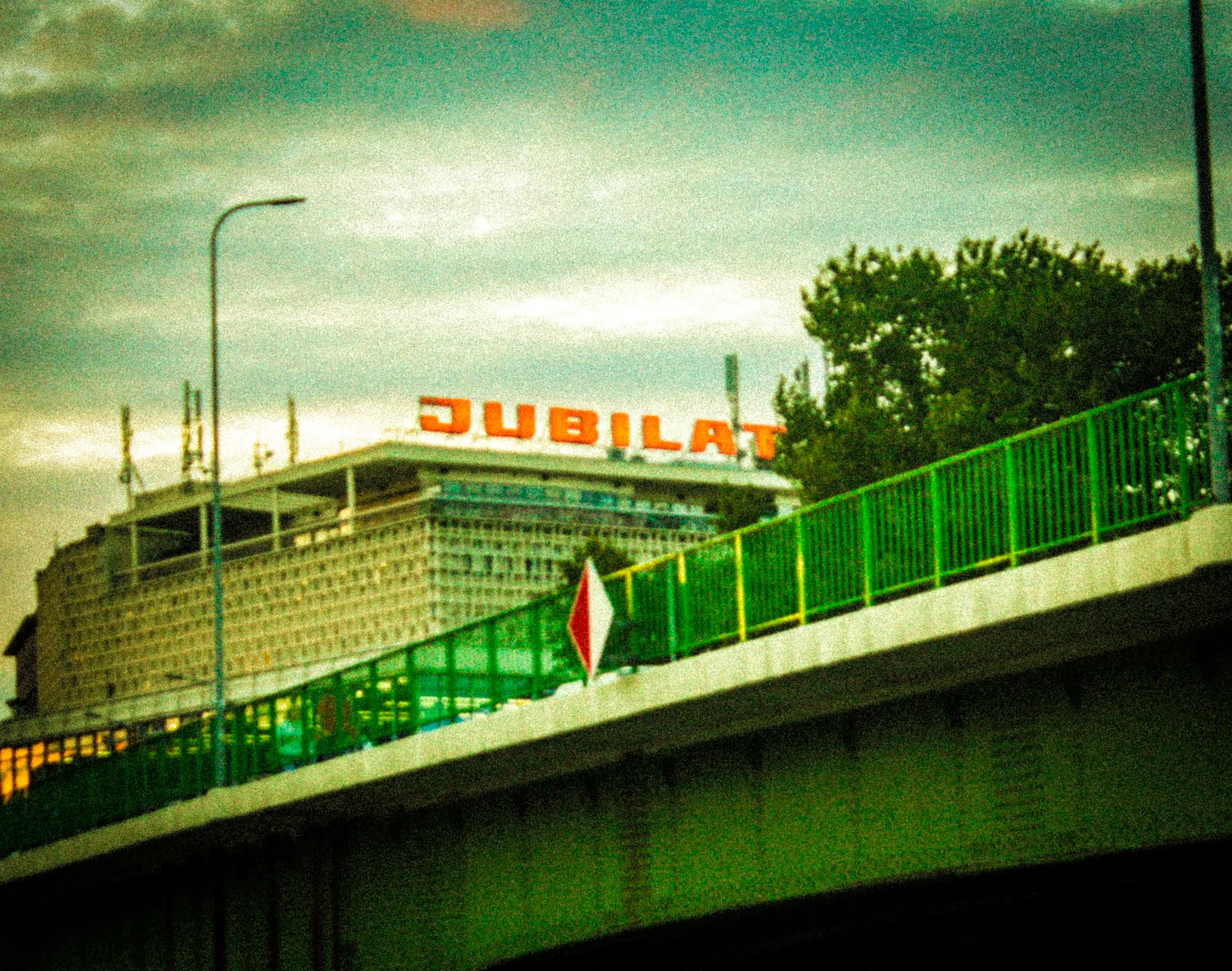 View of a building at night with a large sign spelling 'JUBILAT' in bright orange lights, and a green railing in the foreground.