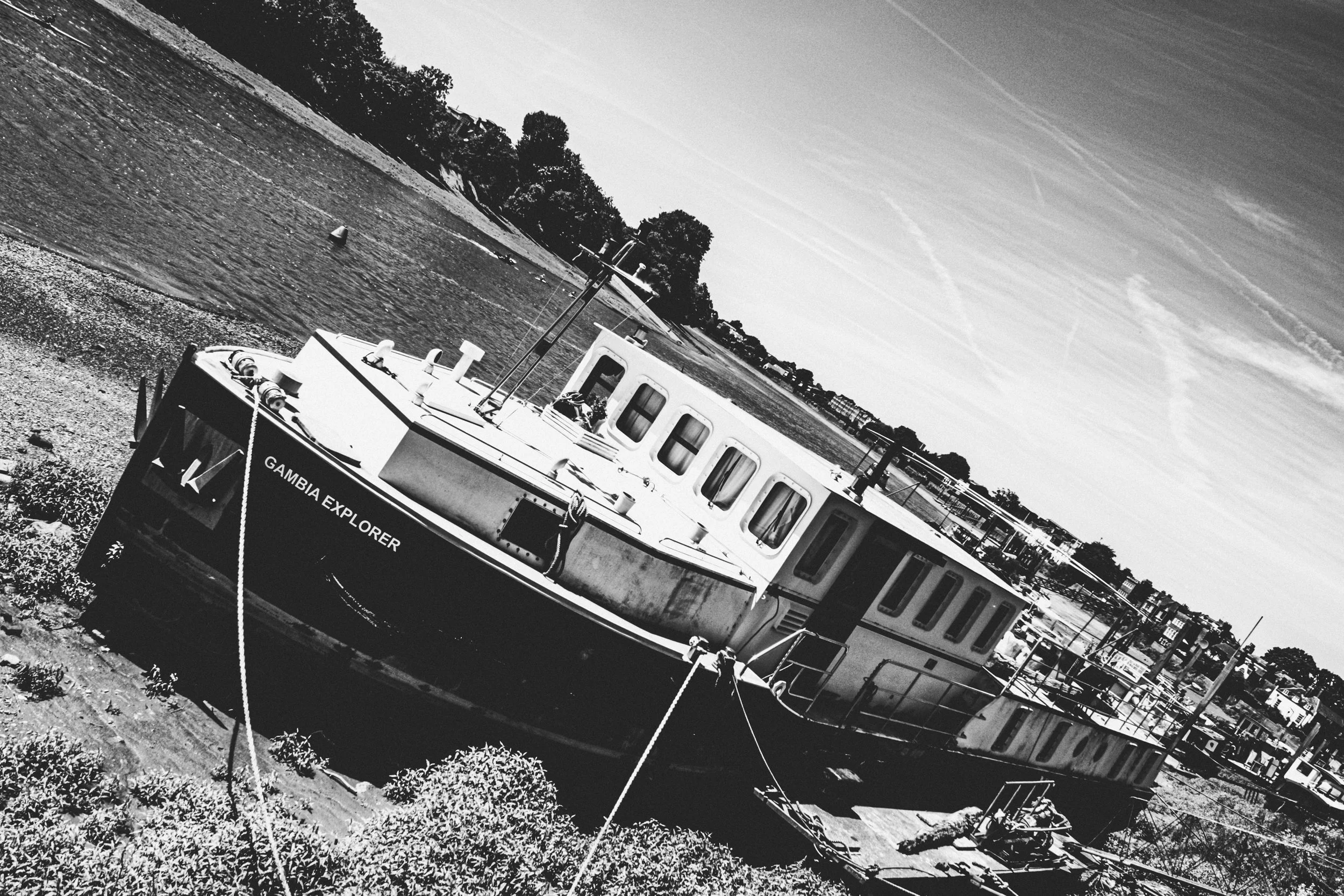 A black and white photograph of a boat named Gambia Explorer docked on a shoreline with trees and buildings in the distance and a sky with streaky clouds.