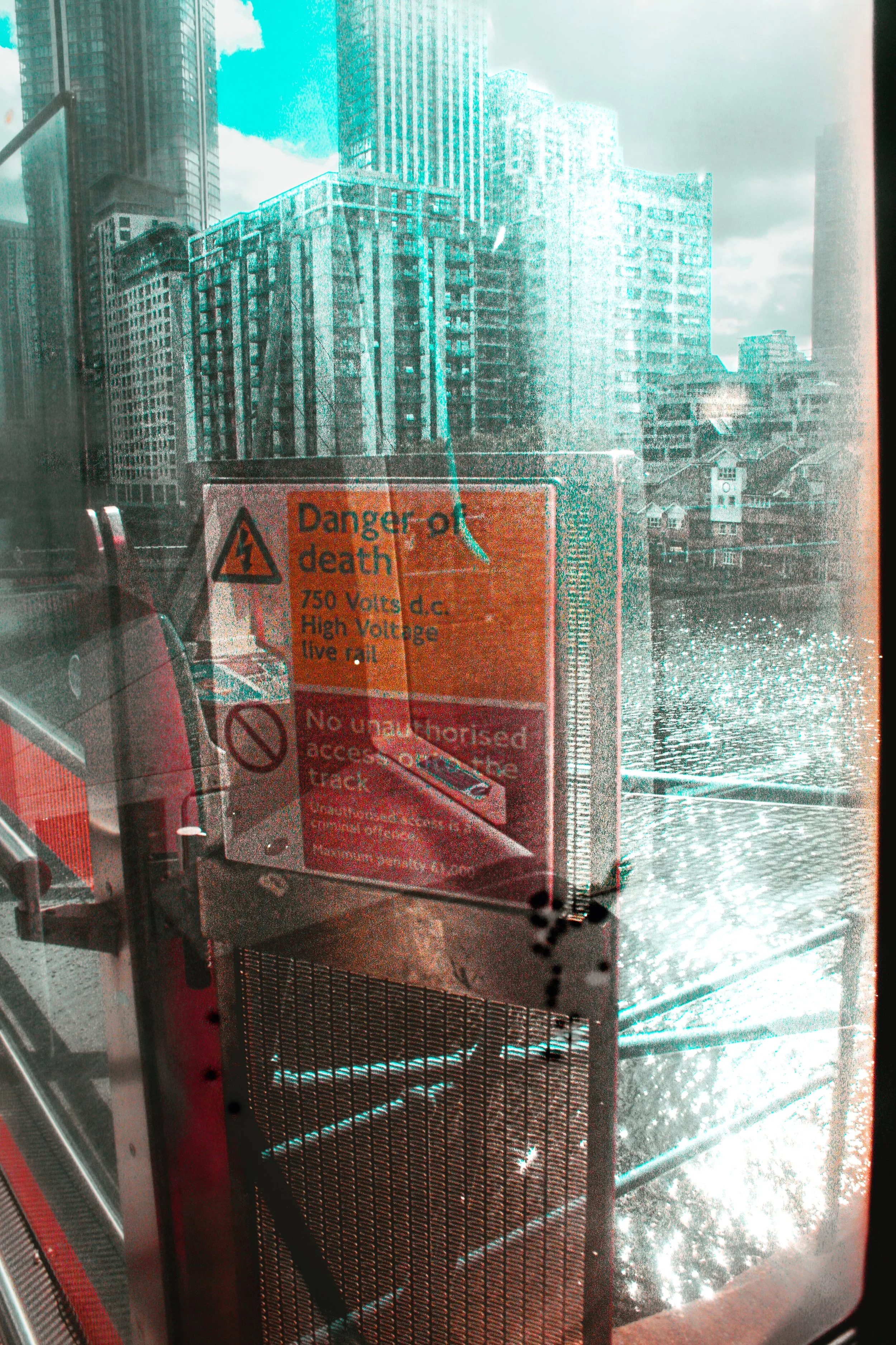 A view of tall buildings through a glass window with caution and warning signs post. The signs indicate danger of death due to high voltage and restricted access.