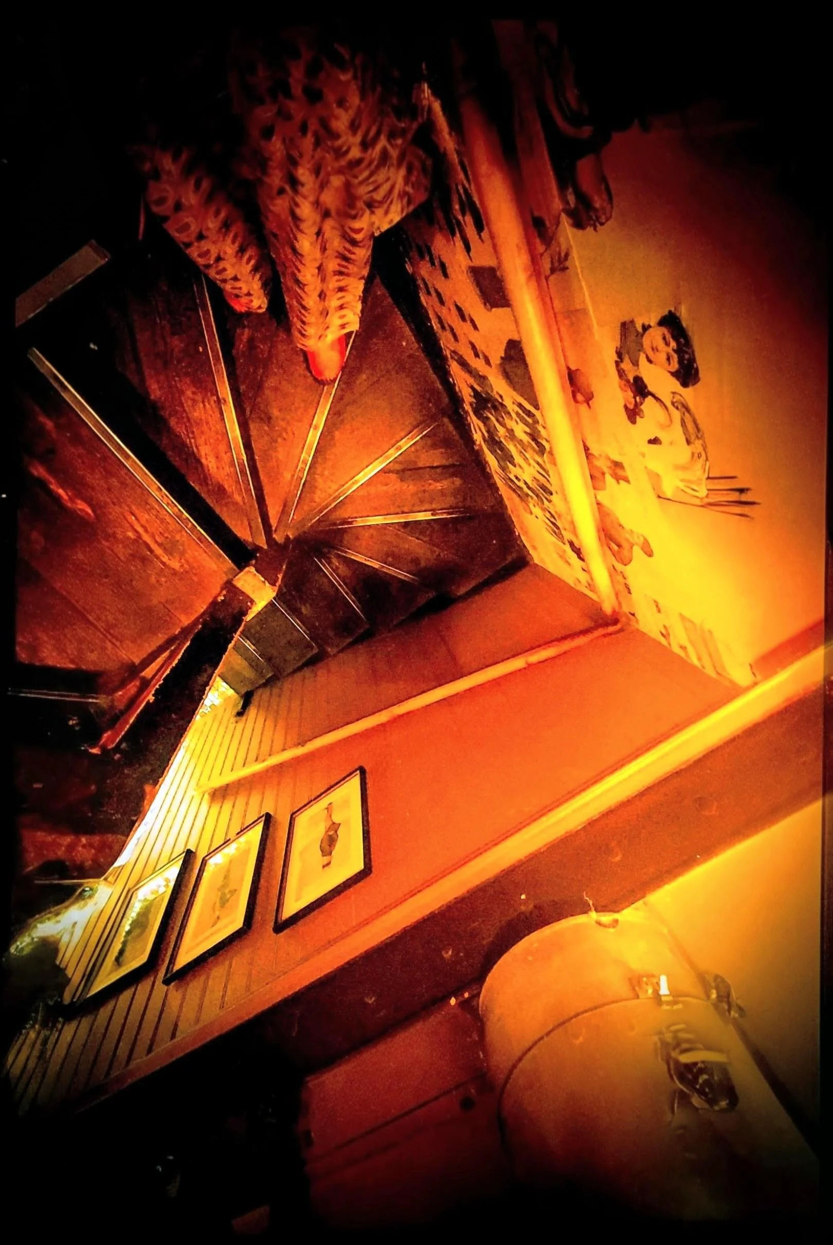 Dimly lit indoor scene with a patterned blanket on a chair, framed pictures on a wood-paneled wall, and a table with a glass and a white object.