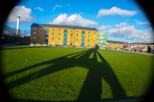 A large yellow, blue, and green building with a grassy area in front and shadows cast by a bridge or structure, with a blue sky and white clouds in the background.