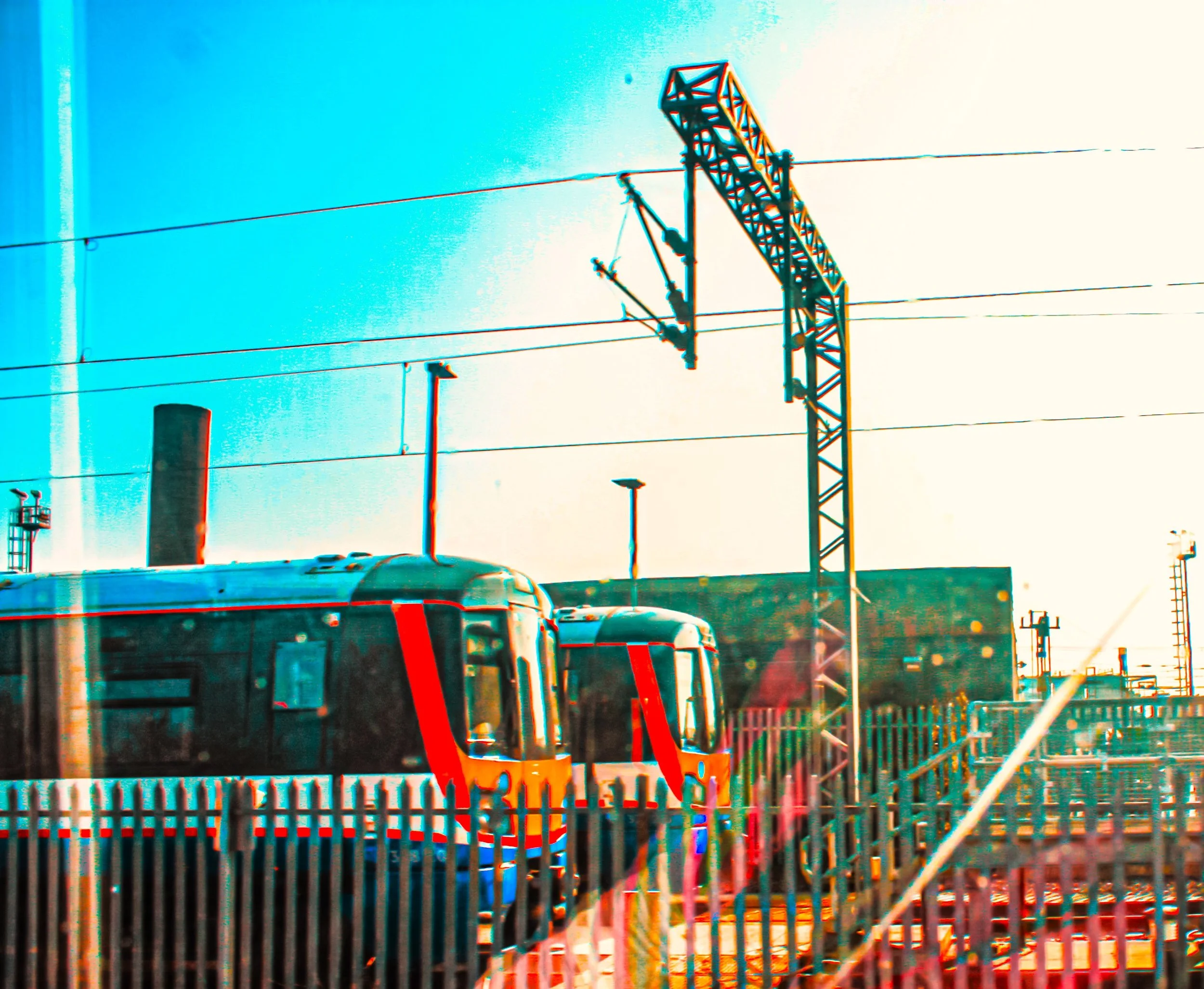 A train at a train station with multiple overhead electrical wires and a fence in the foreground.