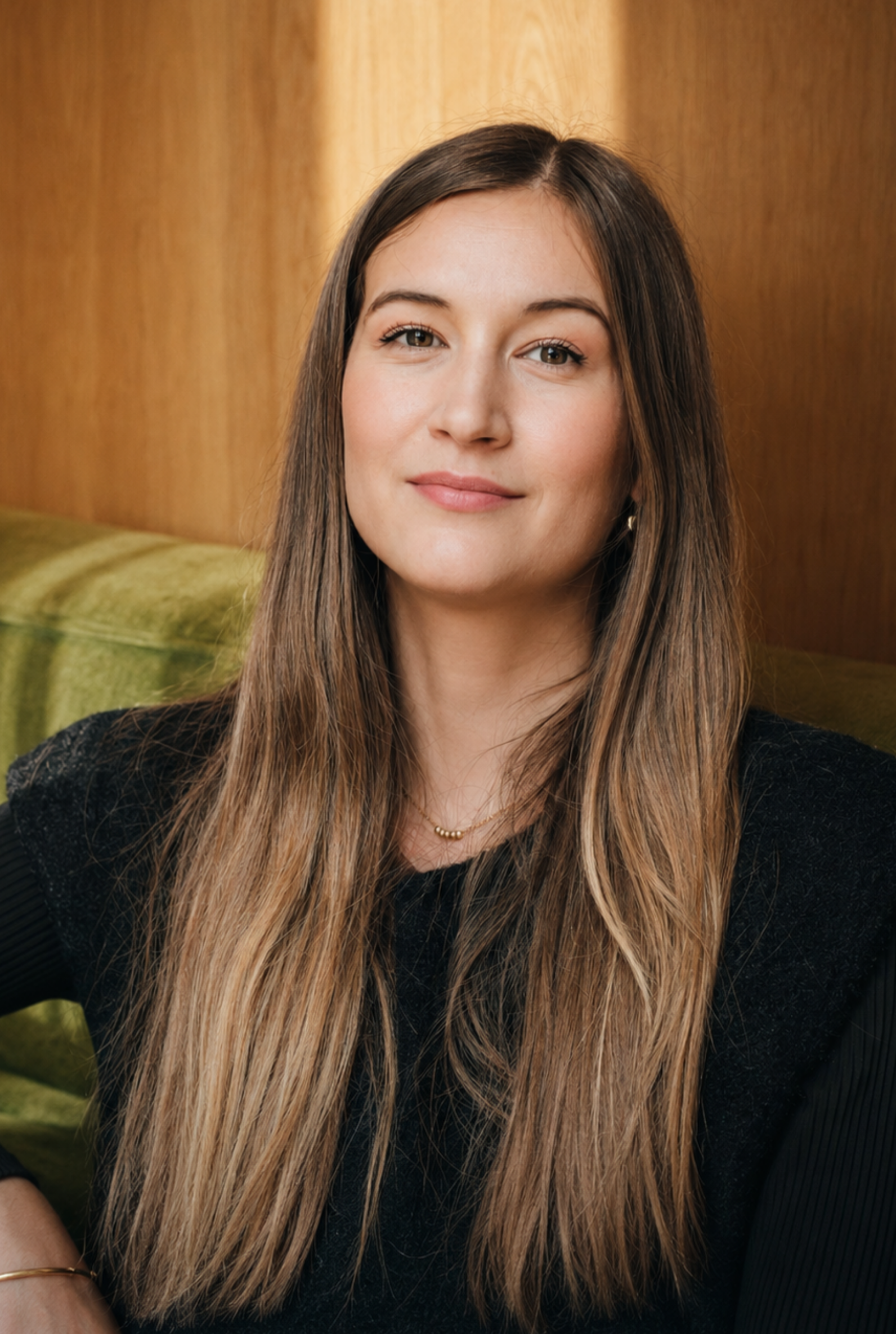 Jeune femme aux longs cheveux bruns et blonds, souriant légèrement, assise dans une pièce avec un fond en bois et une banquette en velours vert olive.