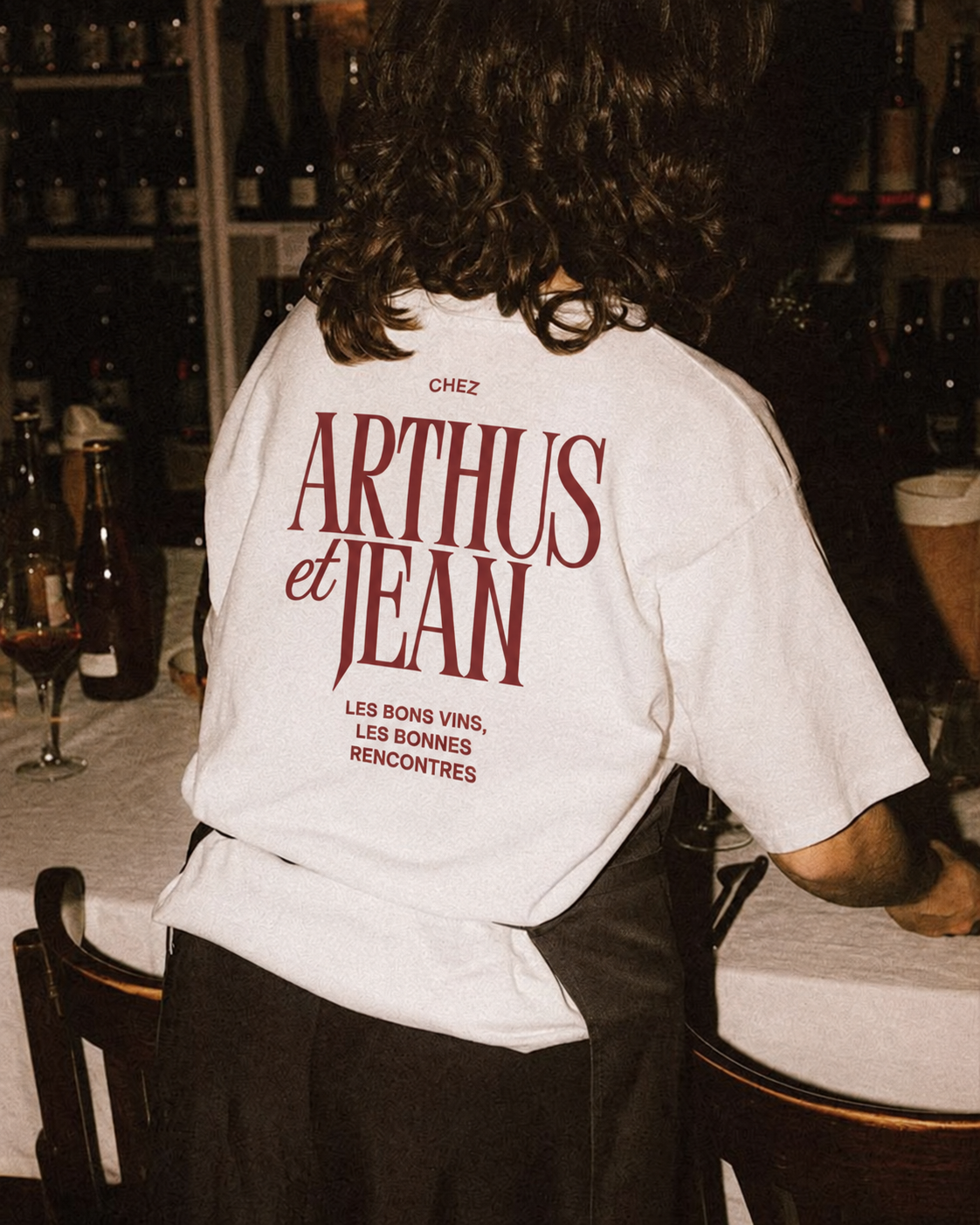 Une personne portant un t-shirt blanc avec le texte 'Chez Arthus et Jean, Les bons vins, Les bonnes rencontres' écrit dessus, dans un environnement probablement un restaurant ou un bar.