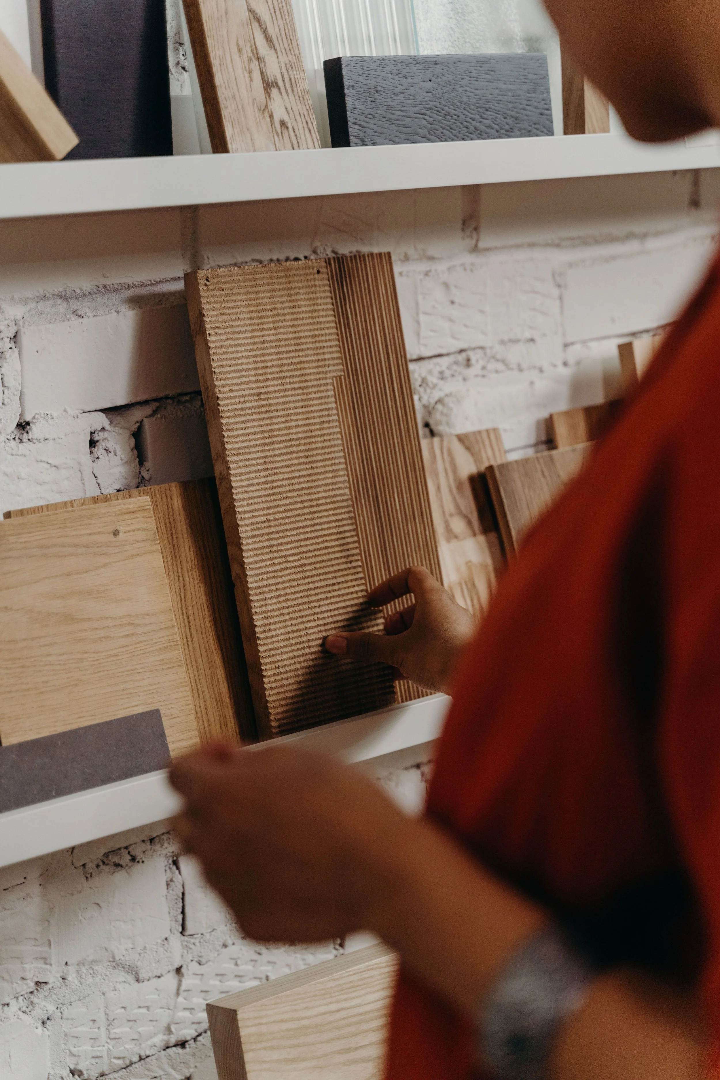 Une personne manipule des échantillons de bois de différentes essences et couleurs exposés sur une étagère blanche, avec un mur en briques blanches en arrière-plan.