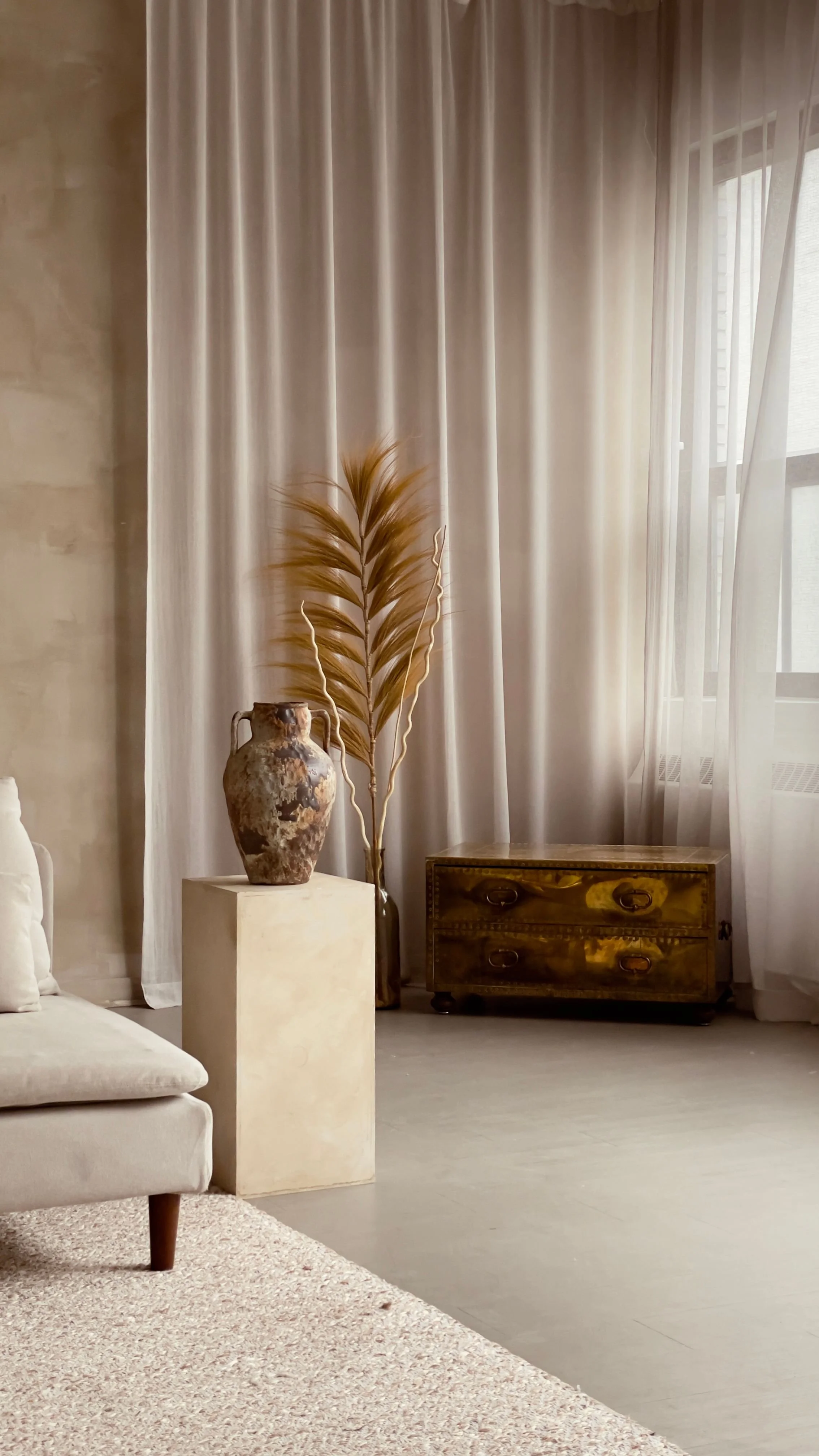 Une pièce élégante avec un rideau blanc, une grande poterie ancienne surun support, une plante séchée, une commode en bois ancienne, un canapé beige en tissu avec un coussin blanc, et un sol en bois clair.