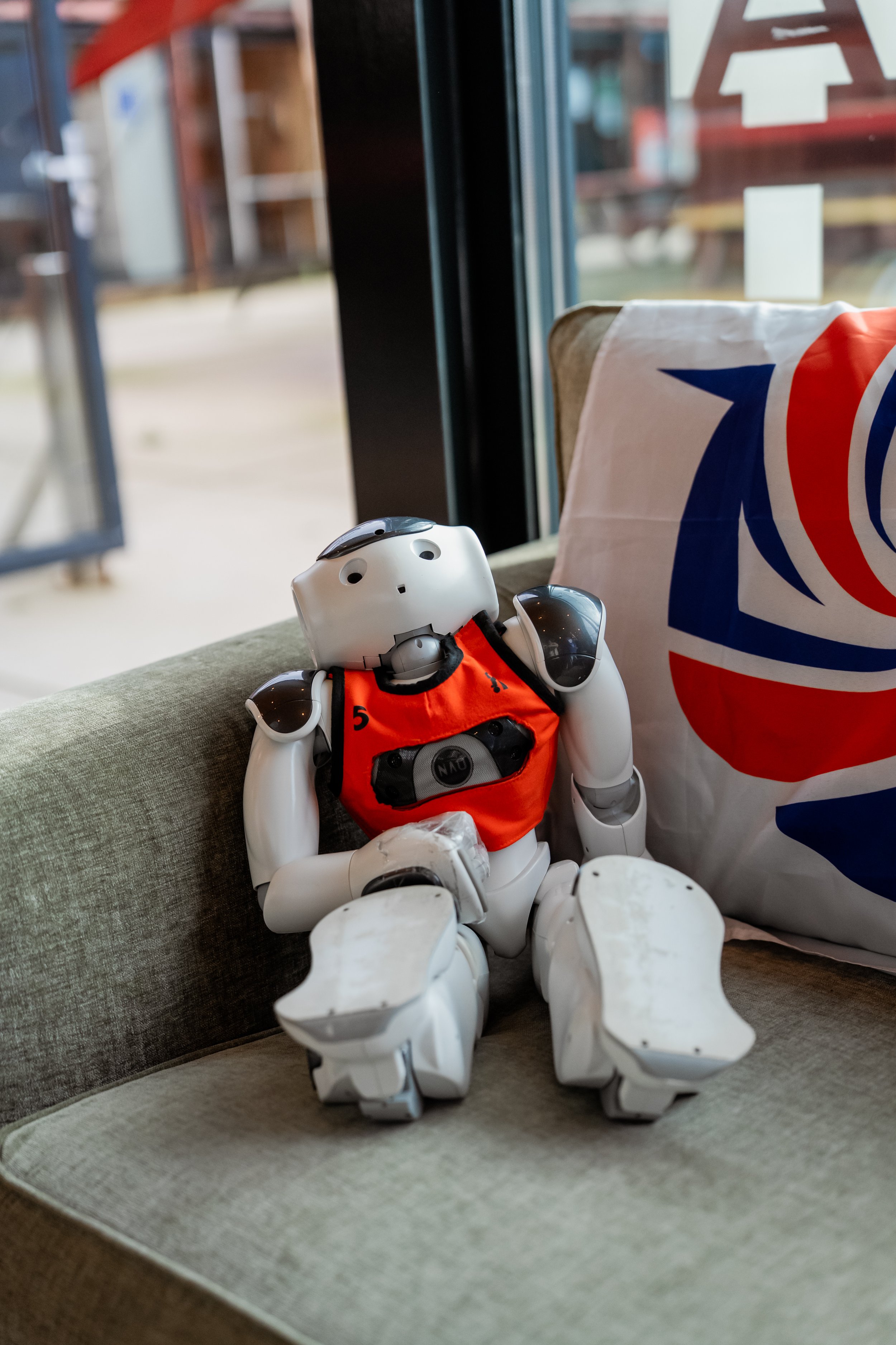 A robot toy with a surprised facial expression, wearing an orange vest, sitting on a green couch next to a shopping bag with a red, white, and blue swirl design.