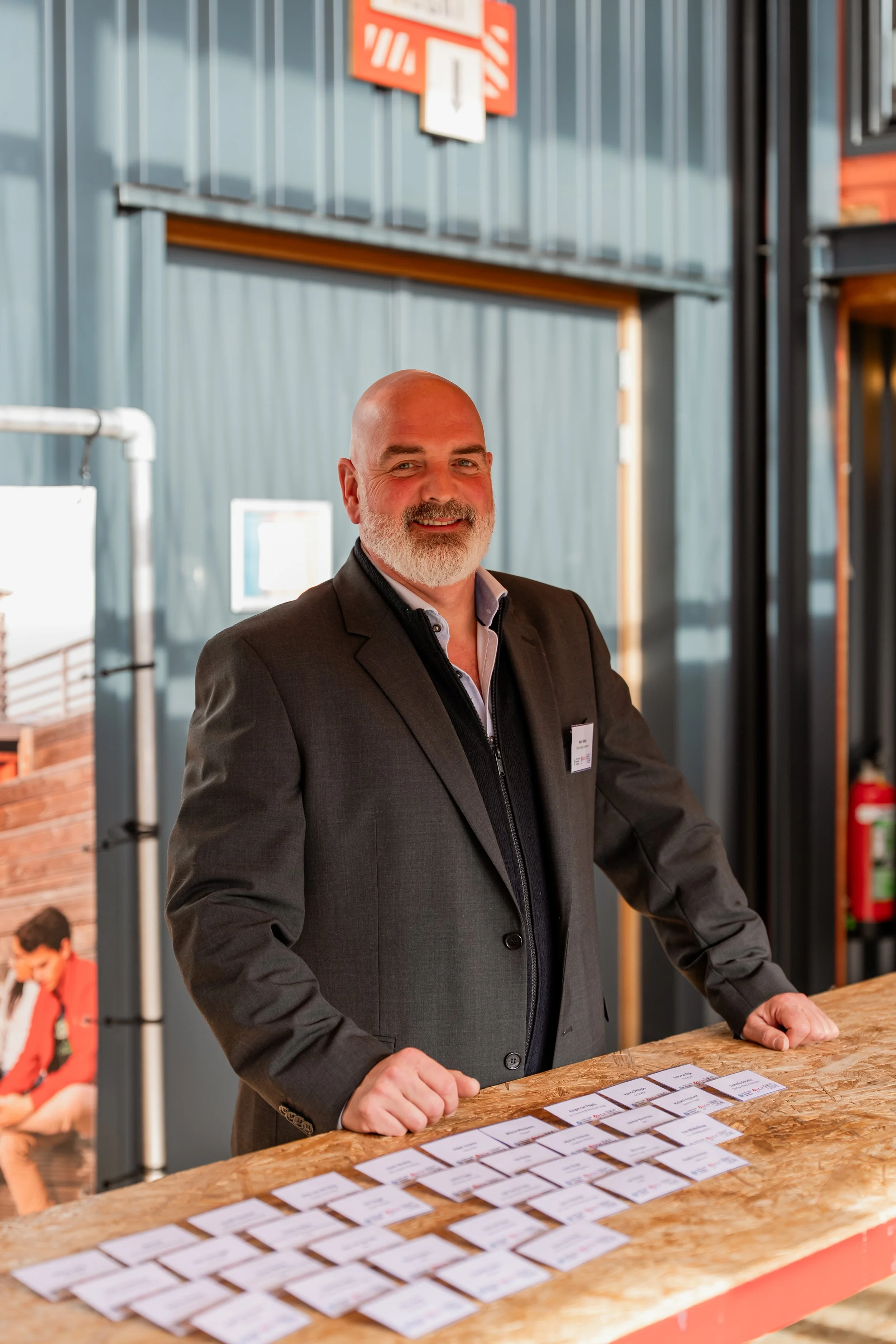 A man in a dark suit and white shirt standing behind a wooden counter with name tags on it, in an industrial-style setting.