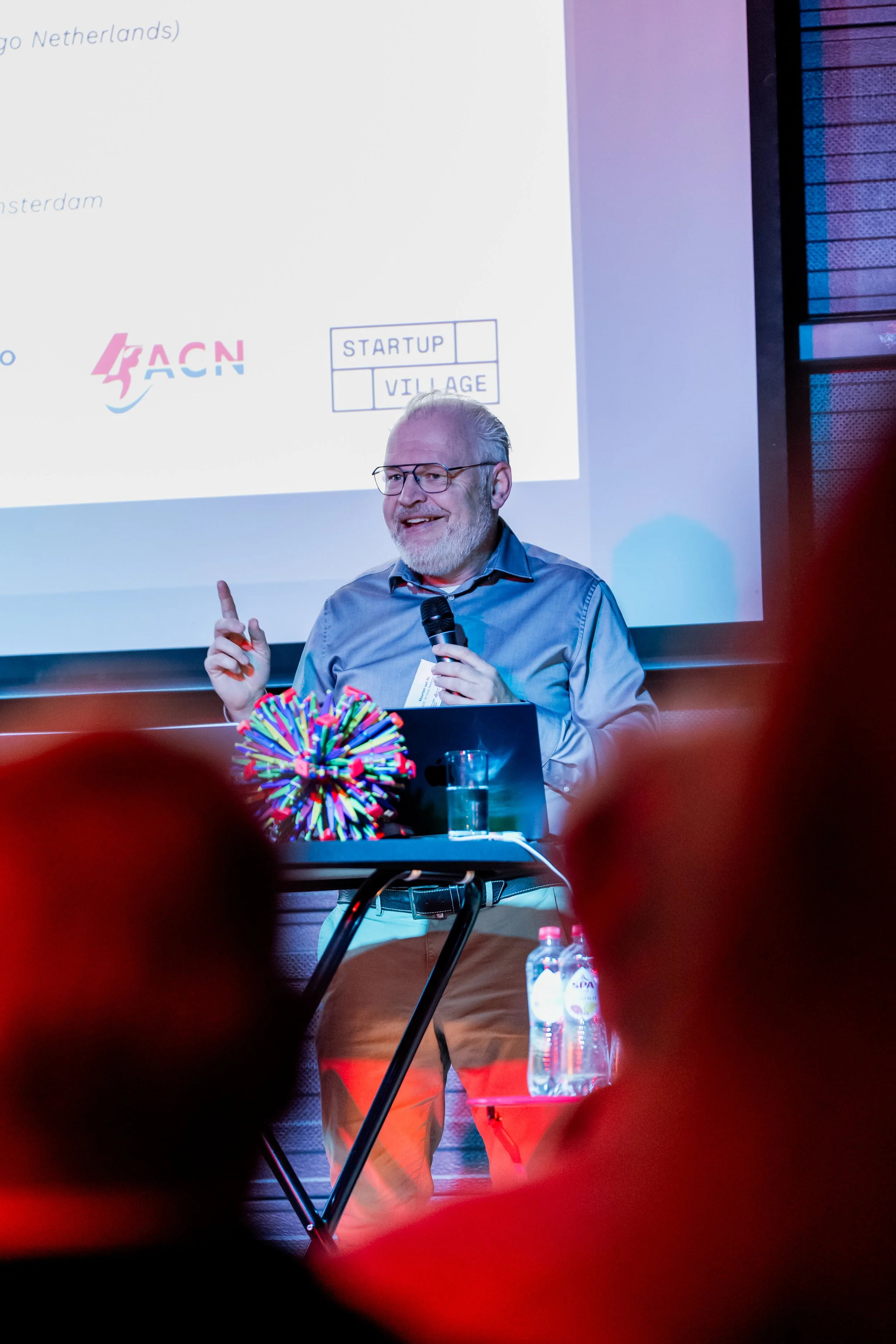 A man with glasses and a beard, holding a microphone, giving a presentation. He is dressed in a blue shirt and beige pants. A colorful decorative object is in the foreground, and water bottles are on a table.