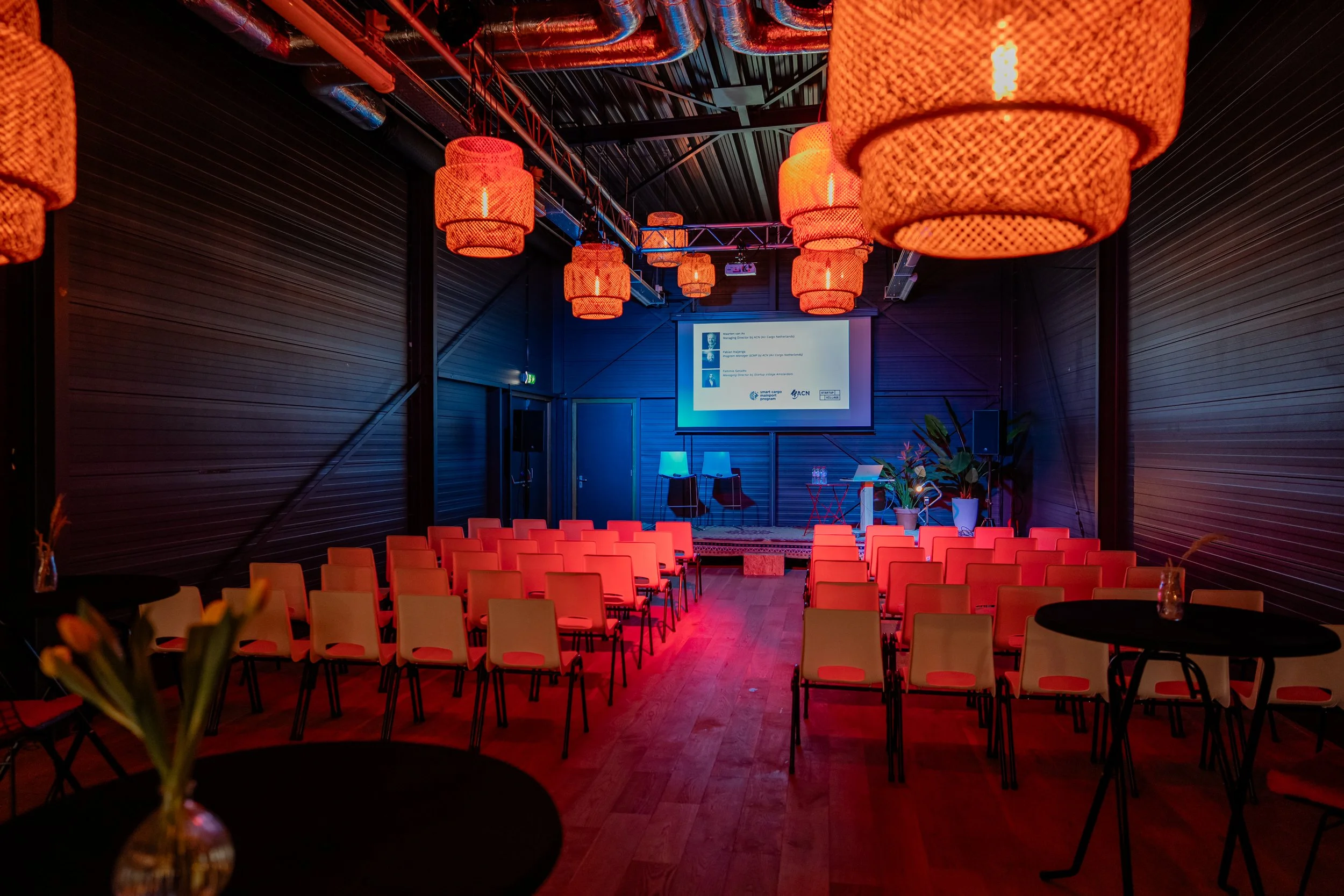 Empty presentation room with rows of orange chairs, a stage with a large screen, hanging orange lamps, and tables with flowers in the foreground.