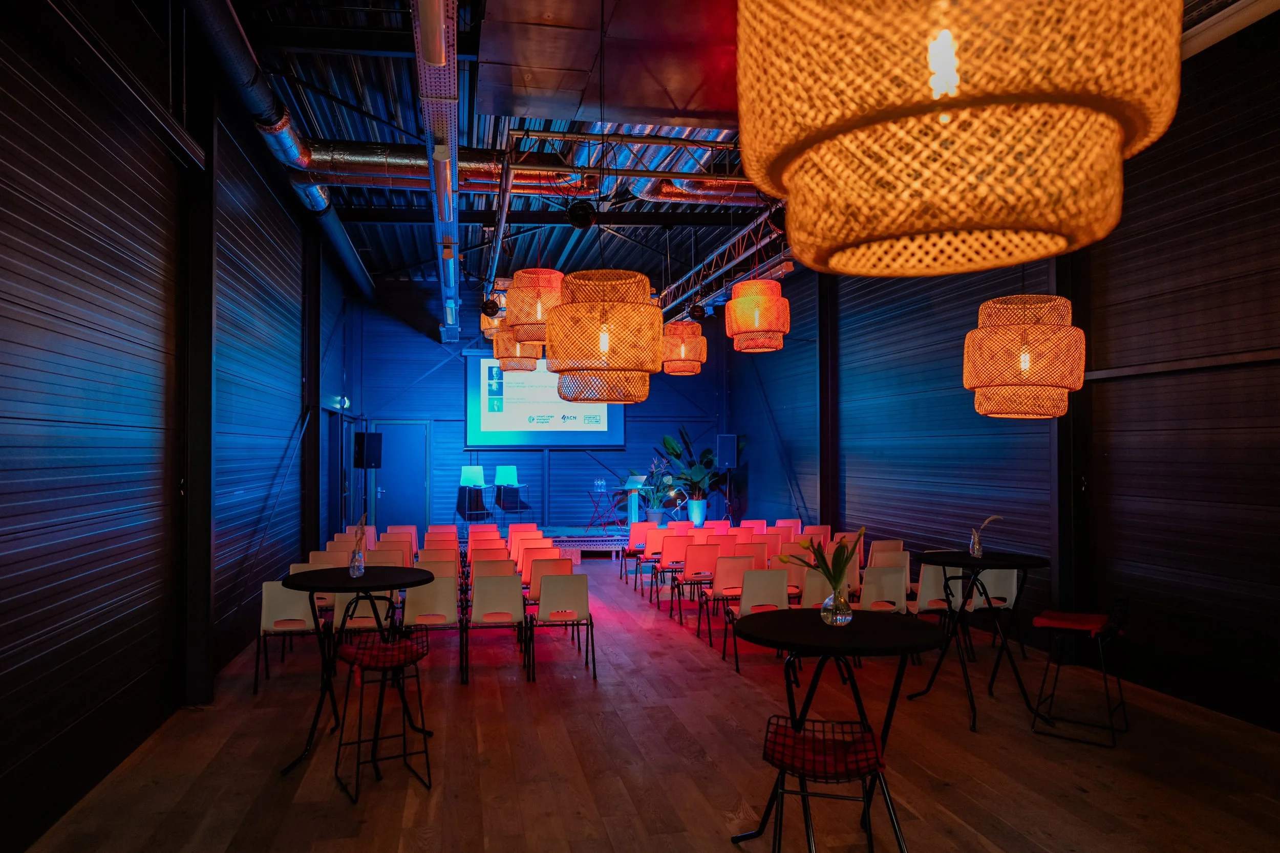 An event space with orange hanging pendant lights and a blue stage screen at the front, set up for a presentation or conference with chairs and tables arranged in rows.