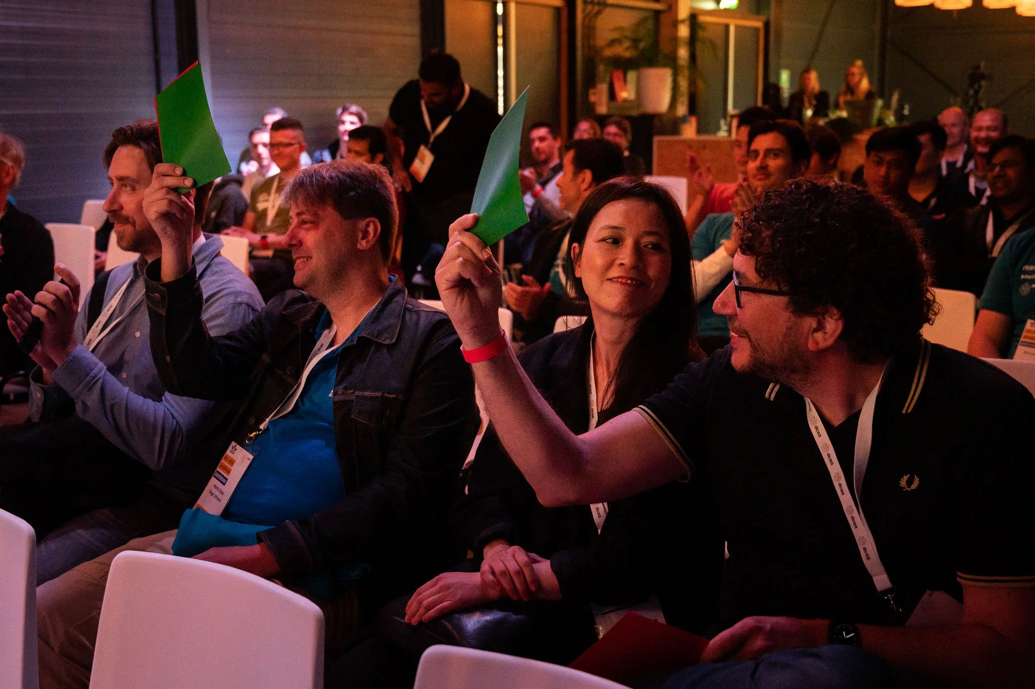 Group of people attending a conference or seminar, some holding green papers, sitting in a dimly lit room with a modern interior.