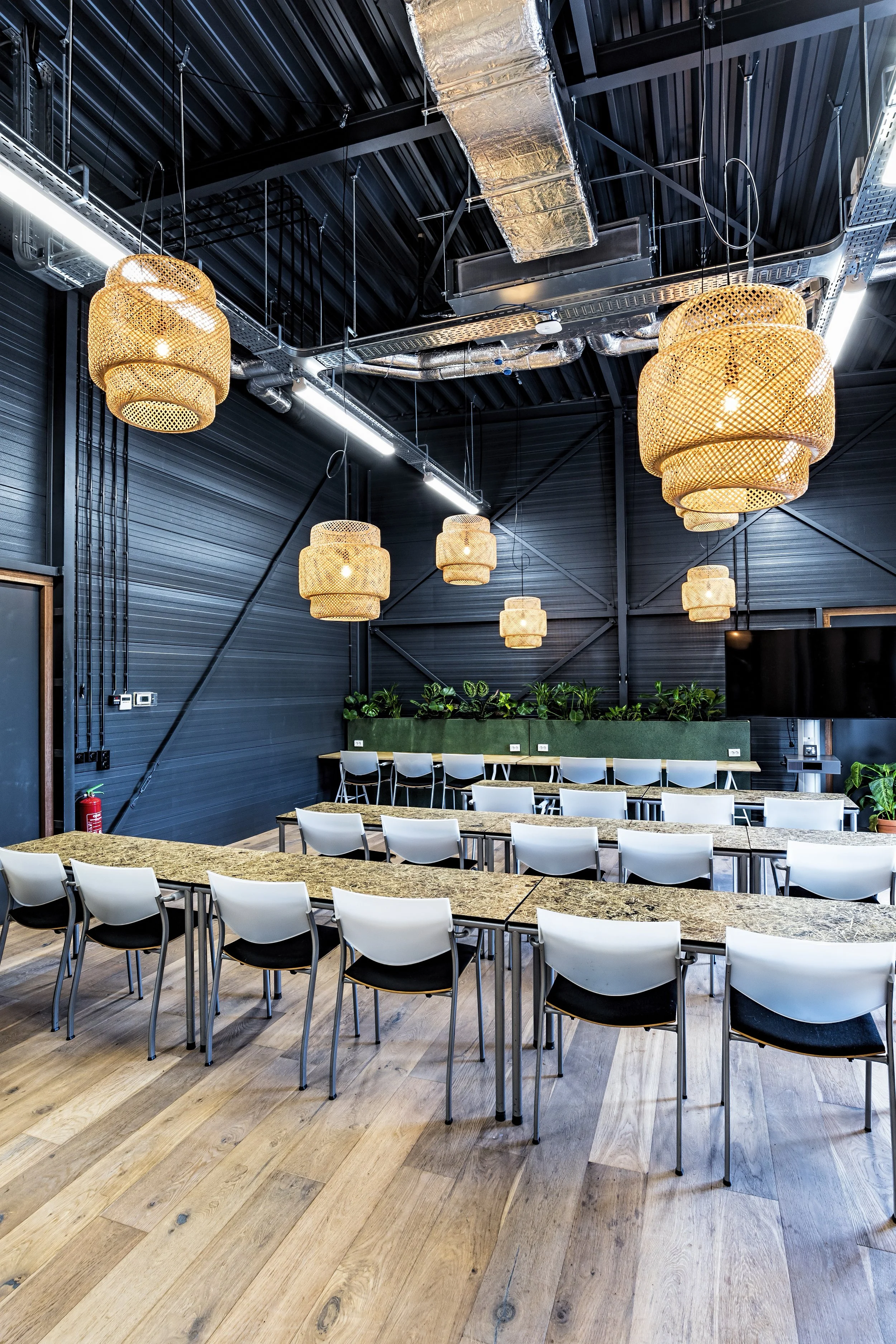 A modern conference room with a curved wooden table surrounded by white chairs, black and gray walls with green plants in the background, and large woven pendant lights hanging from an industrial ceiling with ductwork.