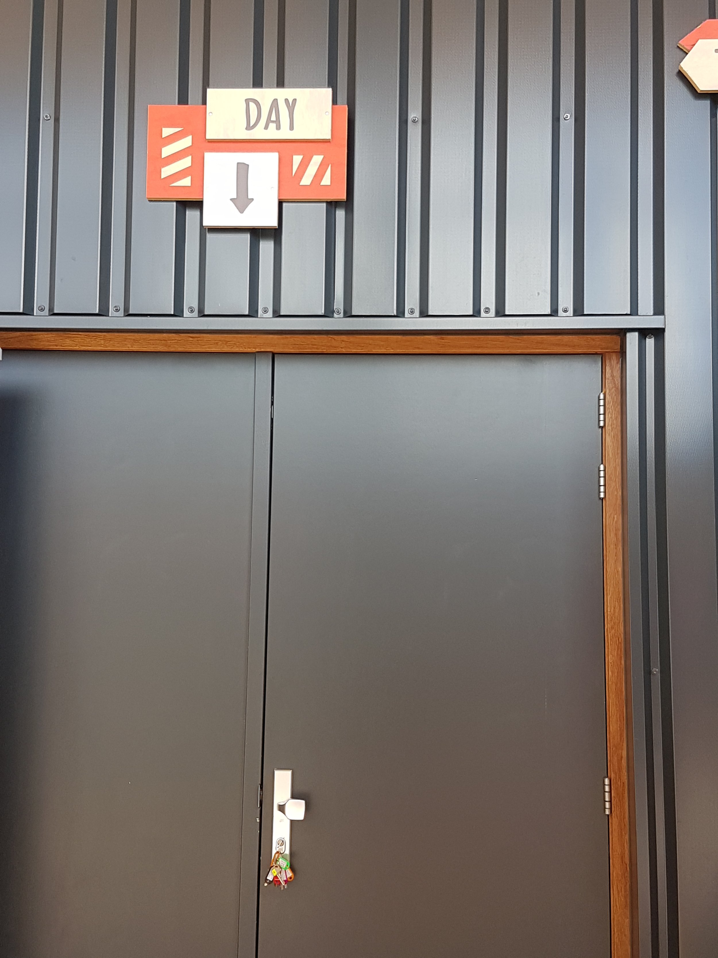A metal door with a silver handle and a set of keys hanging from it. Above the door, there is a decorative sign with the word 'DAY' and an arrow pointing downward. The background features dark gray vertical wall panels with a strip of wood trim along the top of the door.