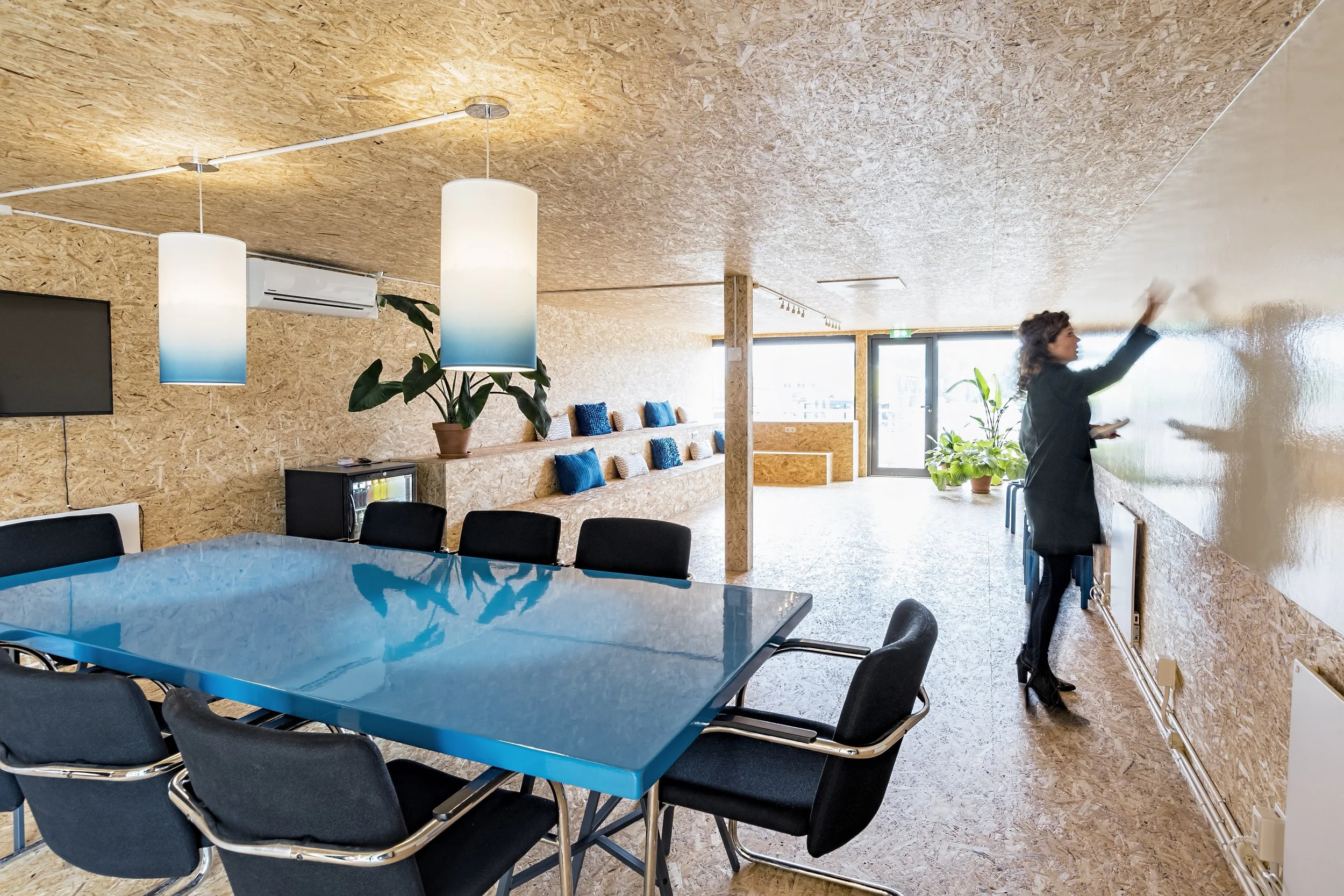 Interior of an office or conference room with a blue table, black chairs, and a woman writing on a whiteboard. The room has wooden walls, hanging pendant lights, and potted plants near large windows.